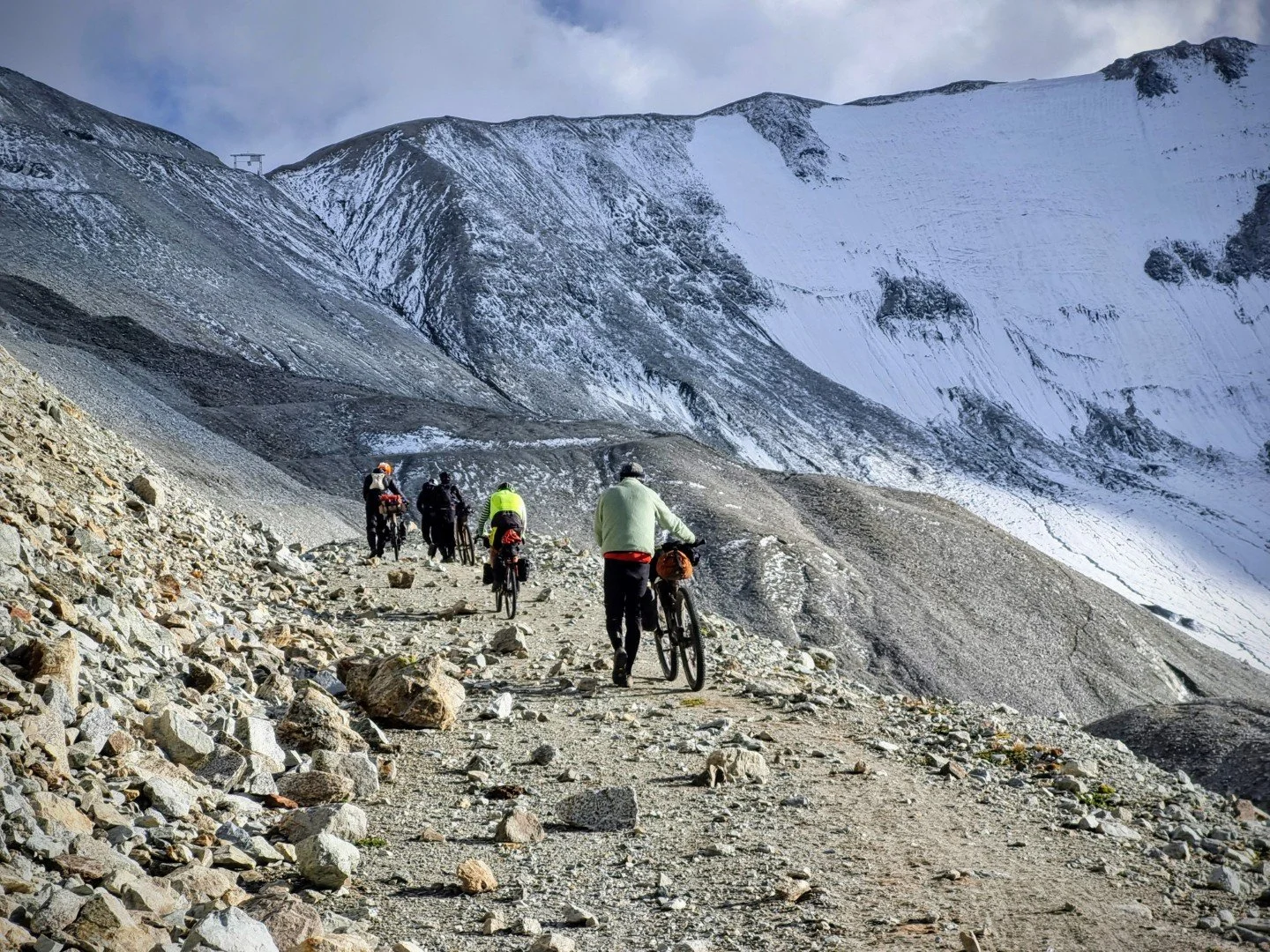 Group of cyclists riding bikepacking bikes on a rocky trail in a snow-covered mountainous landscape.  Heading towards a high mountain pass in Kyrgyzstan on the Silk Road Mountain Race ultra endurance bikepacking event.