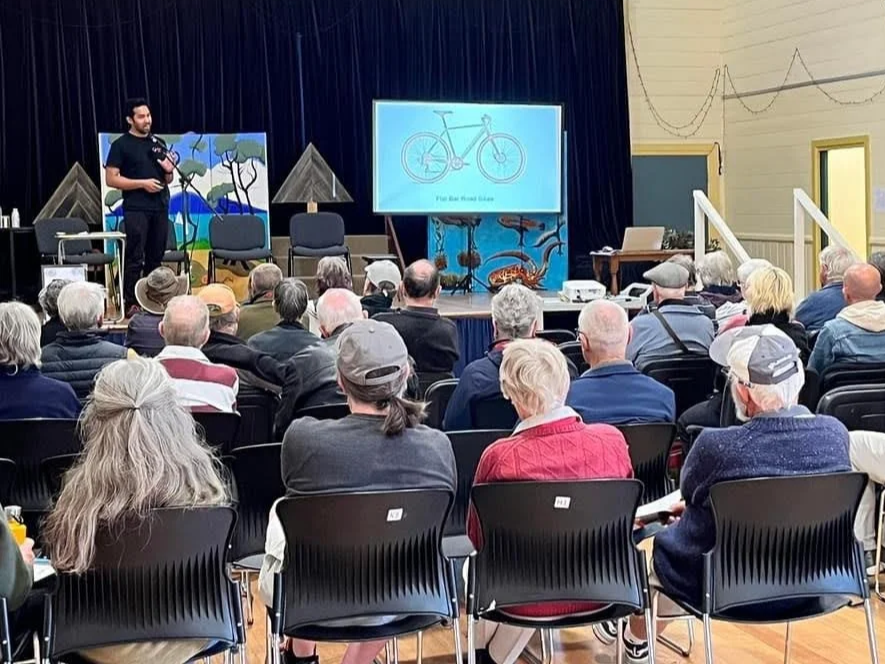 John Paul Lopez Taberdo standing on stage with a microphone, teaching about e-bikes in front of an audience of mostly older adults seated in black chairs, in a room with colorful wall art and decorations.