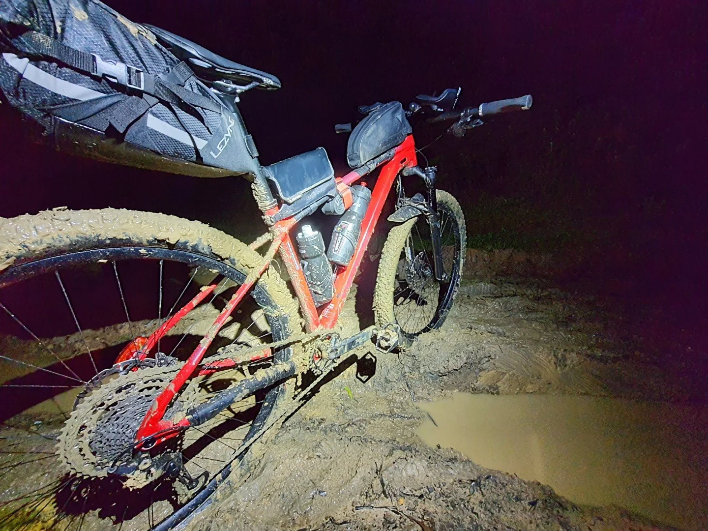 A red mountain bike covered in mud, resting on a wet, muddy trail at night, with a water bottle attached to the frame and a small black saddlebag mounted behind the seat.