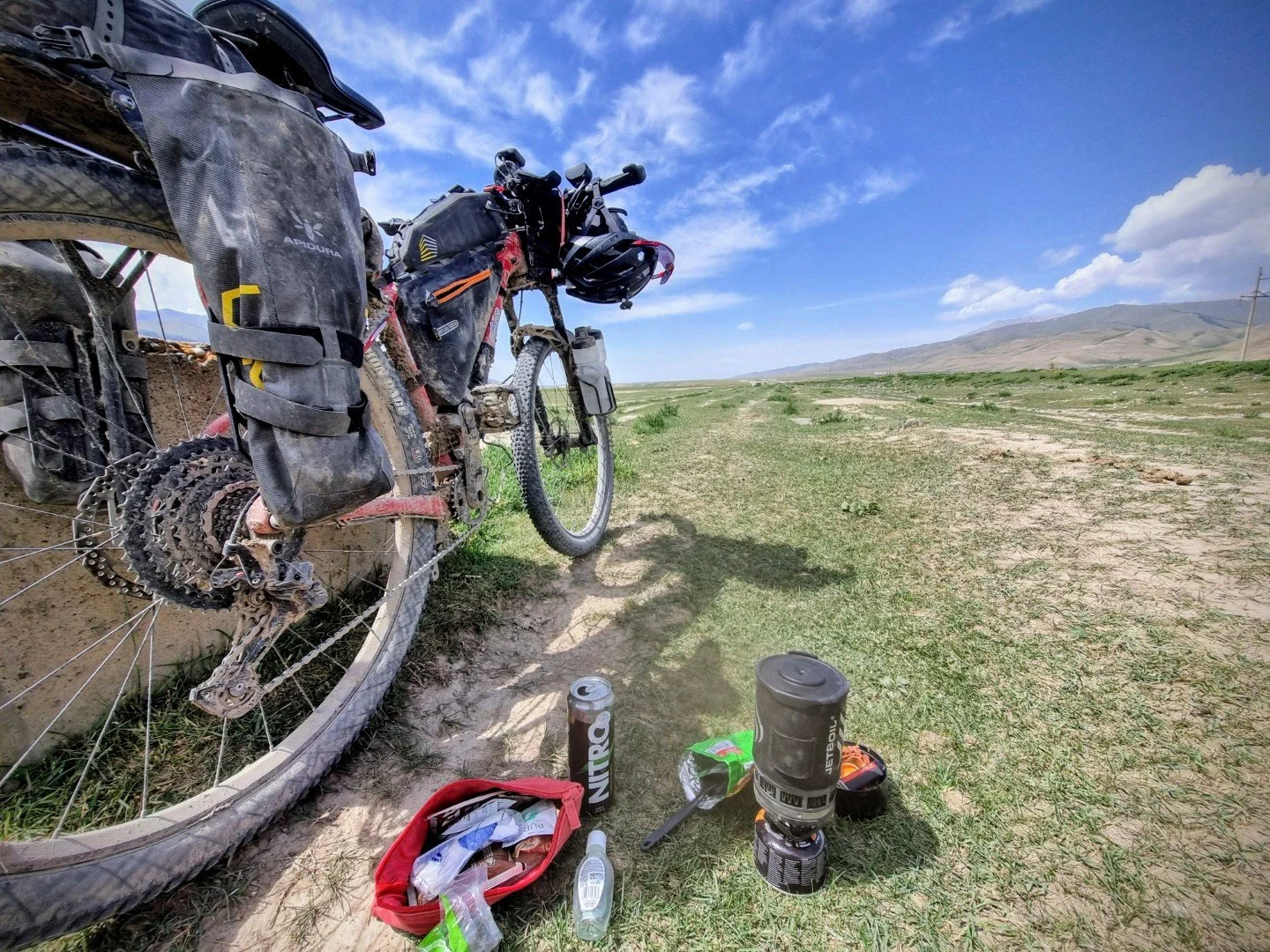 A heavily loaded mountain bike resting on a dirt trail in a grassy open landscape with mountains in the distance under a partly cloudy sky, surrounded by various gear and supplies including a red bag, water bottles, and a portable stove.  Kyrgyzstan.