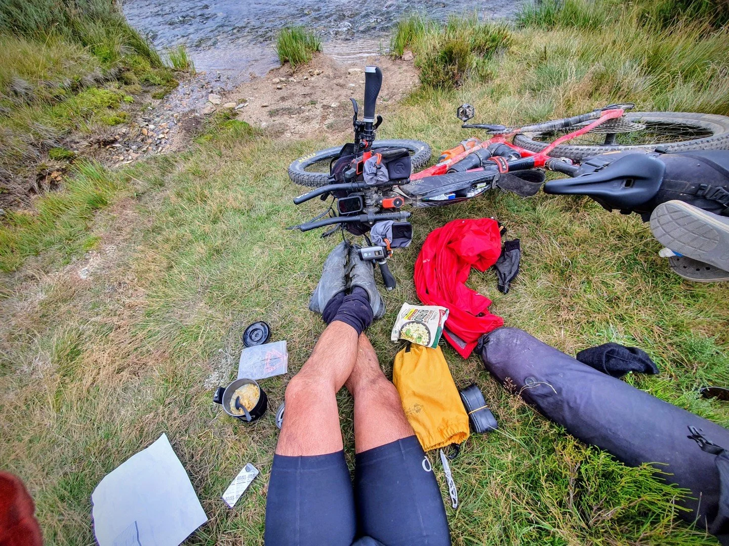 A person sitting on grass at the edge of a lake with a mountain bike nearby, surrounded by gear, food, and supplies.