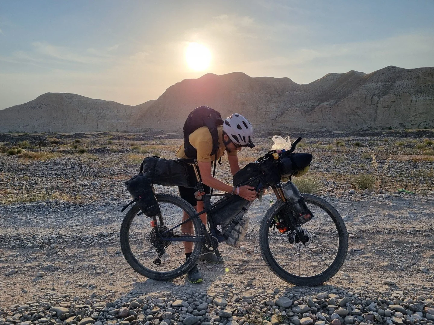 A Silk Road Mountain Race rider in Kyrgyzstan in a yellow shirt, helmet, and sunglasses, with a backpack, leaning over their black mountain bike in a rocky desert landscape with mountains in the background and the sun setting behind the mountains.