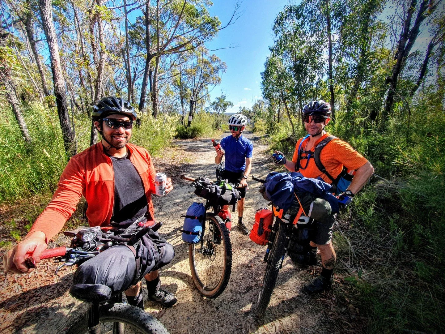 Three men with mountain bikes taking a break on a dirt trail in a wooded area, smiling and holding drinks, wearing helmets and outdoor cycling gear.