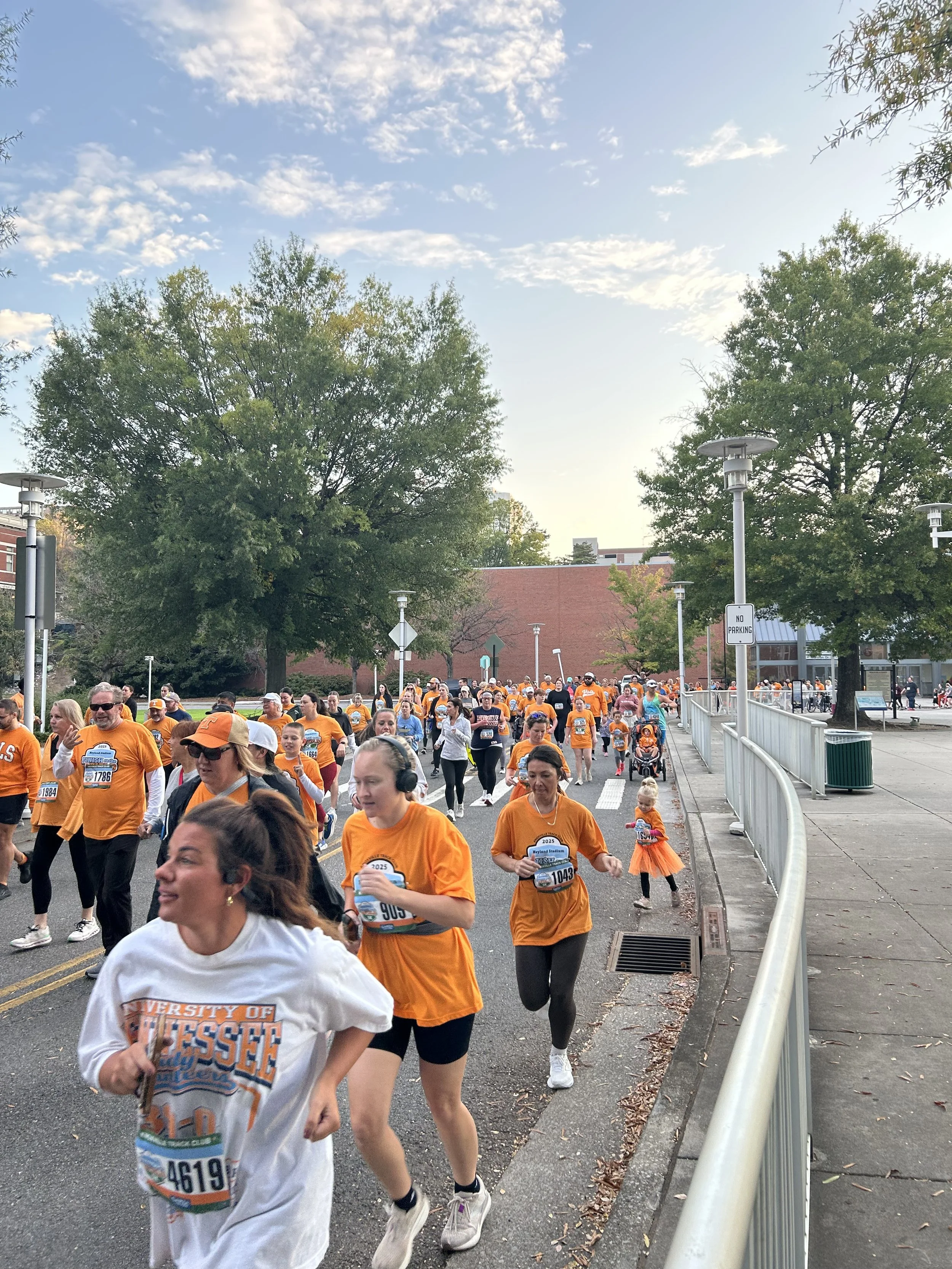 A group of people participating in a race or marathon on a city street, many wearing orange shirts, with some runners near the camera and others farther back, with trees and a brick building in the background.