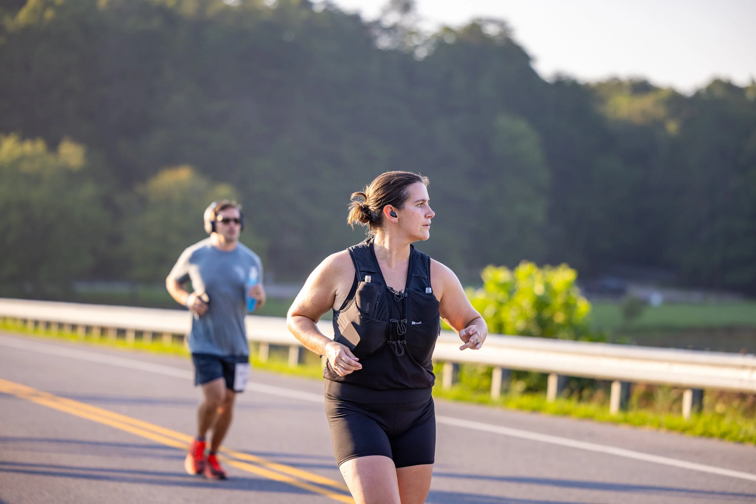 A woman running on a paved road with a man jogging behind her, both wearing athletic gear and headphones, countryside scenery in the background.