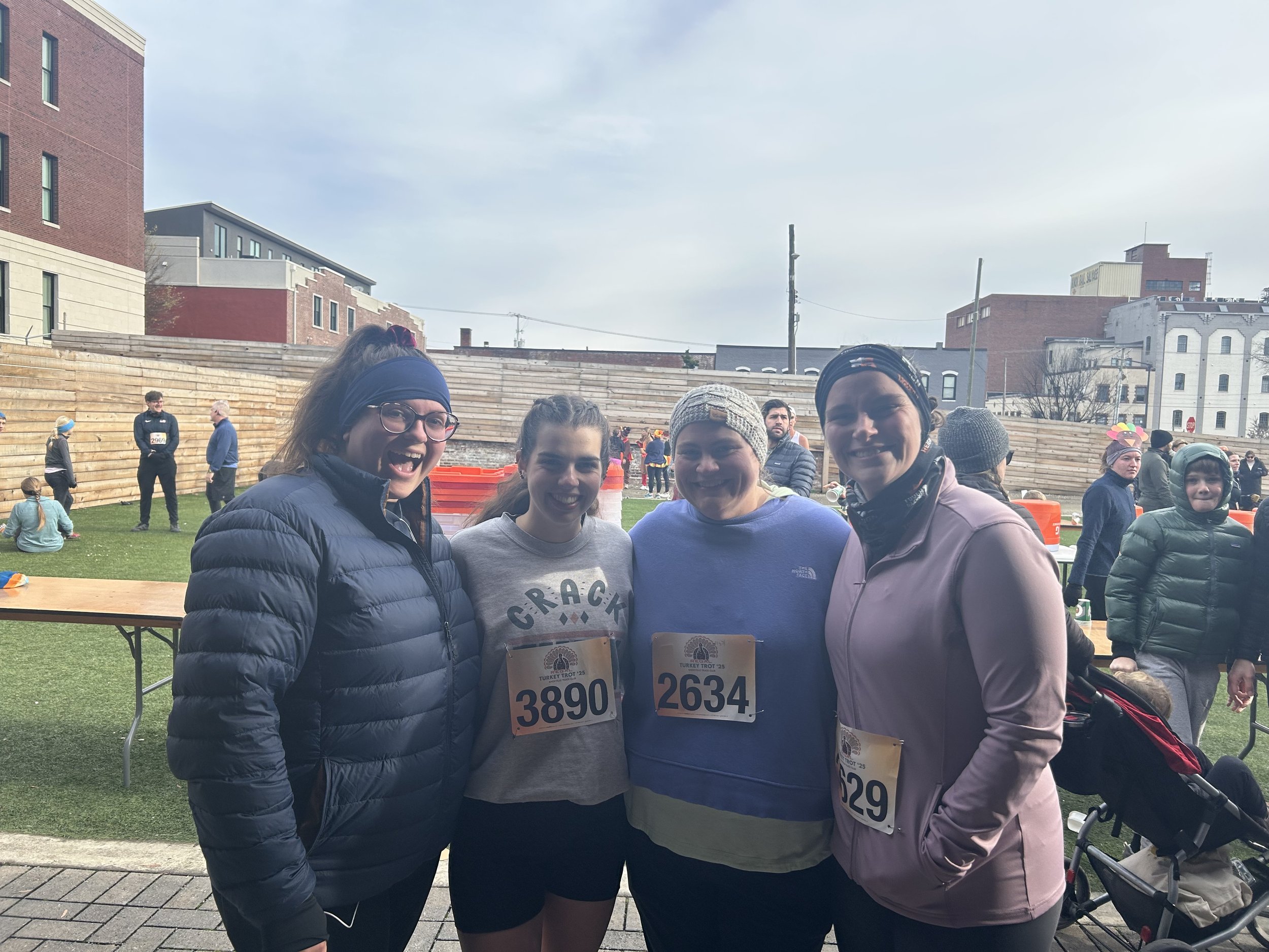 Four women (Run Club Members!) in athletic clothing and numbered bibs smiling at a race event outdoors, with other participants and spectators in the background.