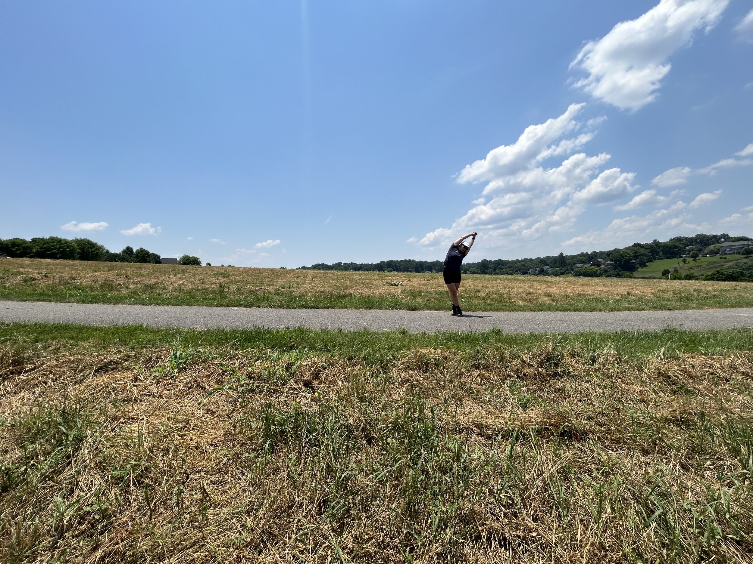 a runner stretches on a path through a field with a large blue sky above