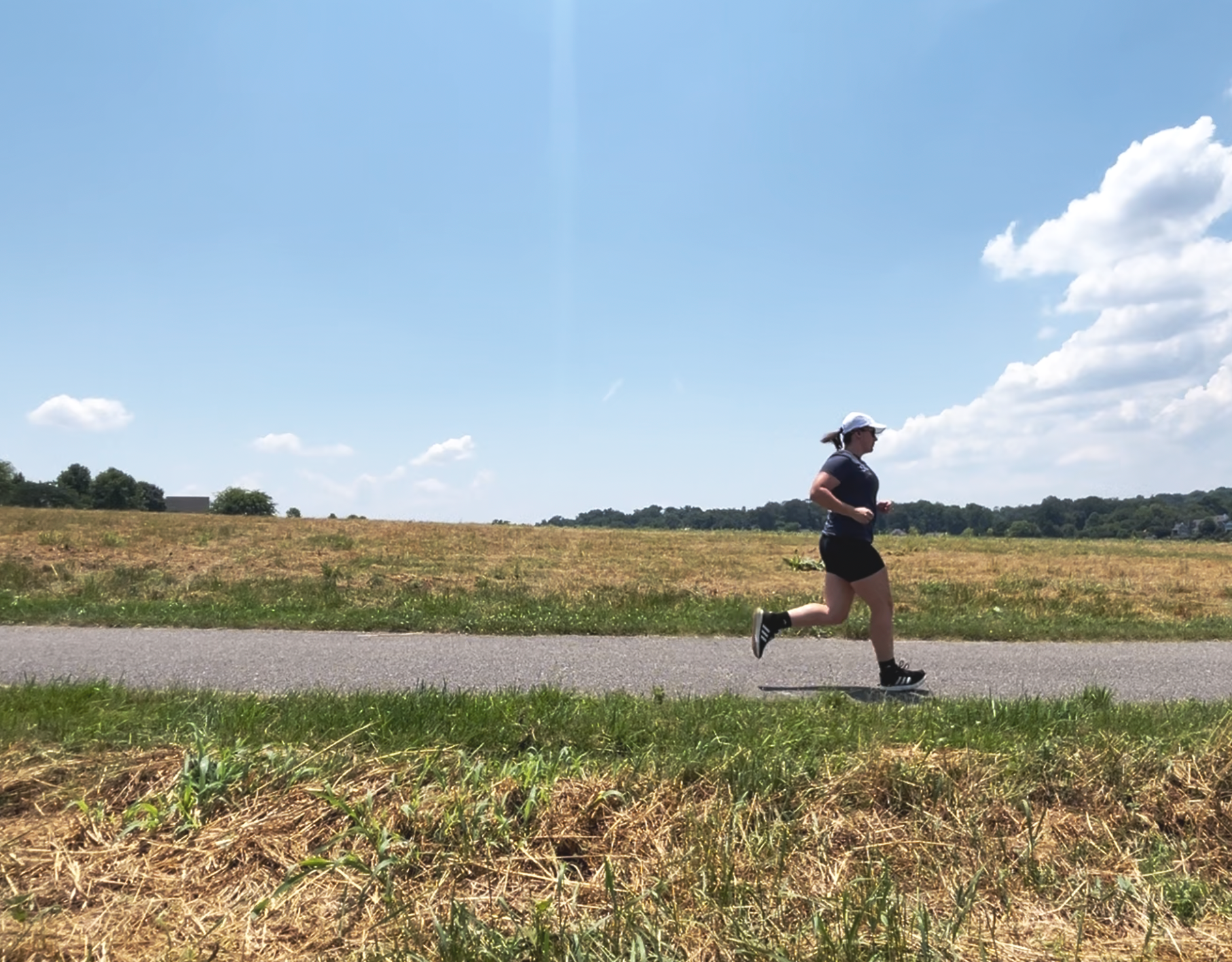 A woman jogging on a paved trail in a rural area under a blue sky with scattered clouds.