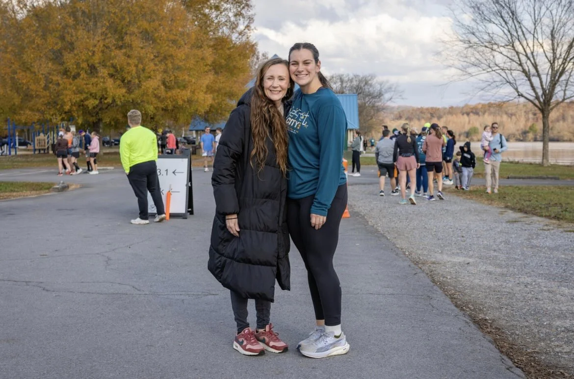 BK and her coach, Mary, smiling together outdoors before a race, with a group of people in the background near trees with autumn leaves.