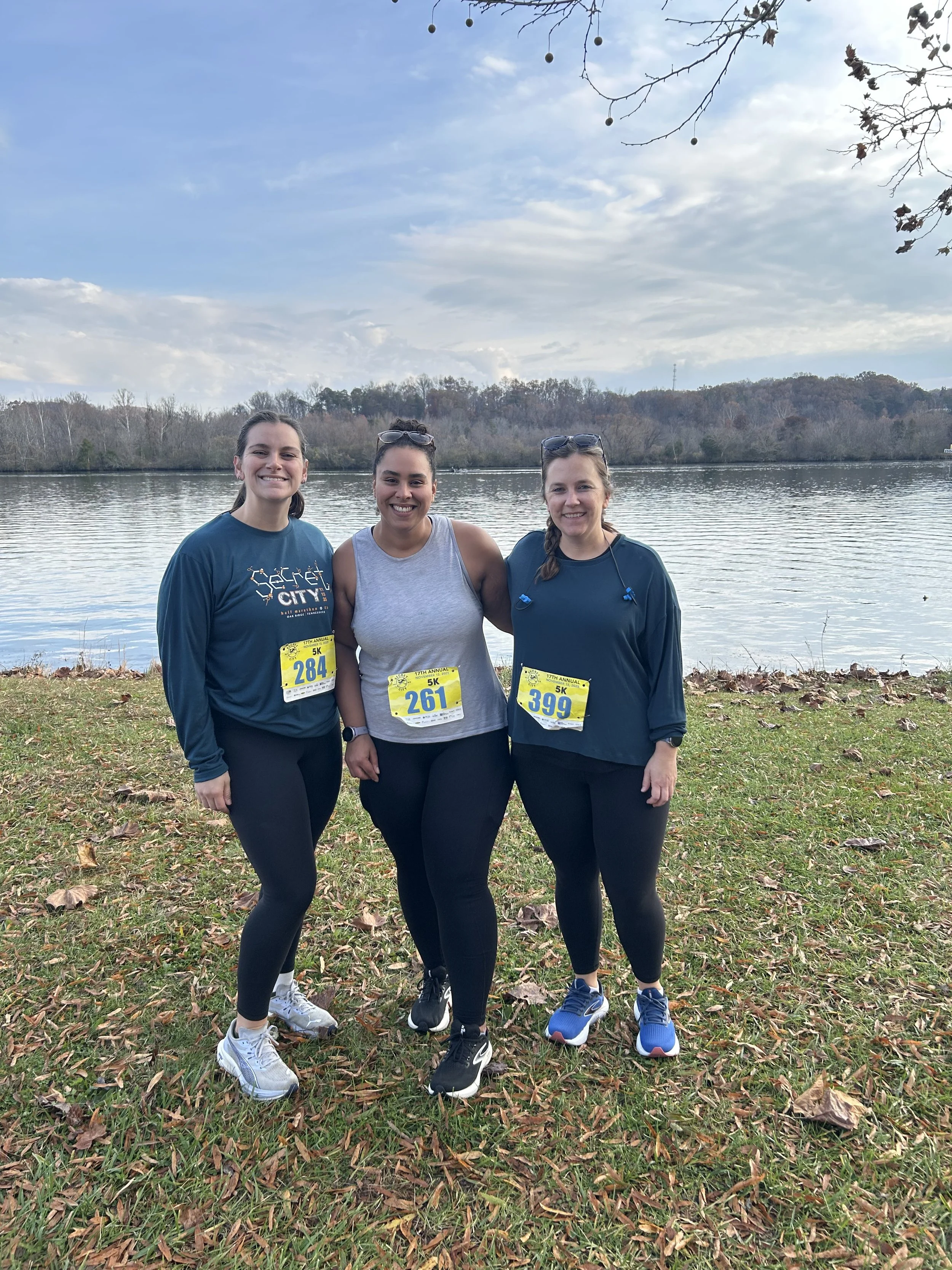 Three women (Run Club Members!) standing outdoors near a lake with a scenic background, smiling, wearing running clothes, and race bibs.