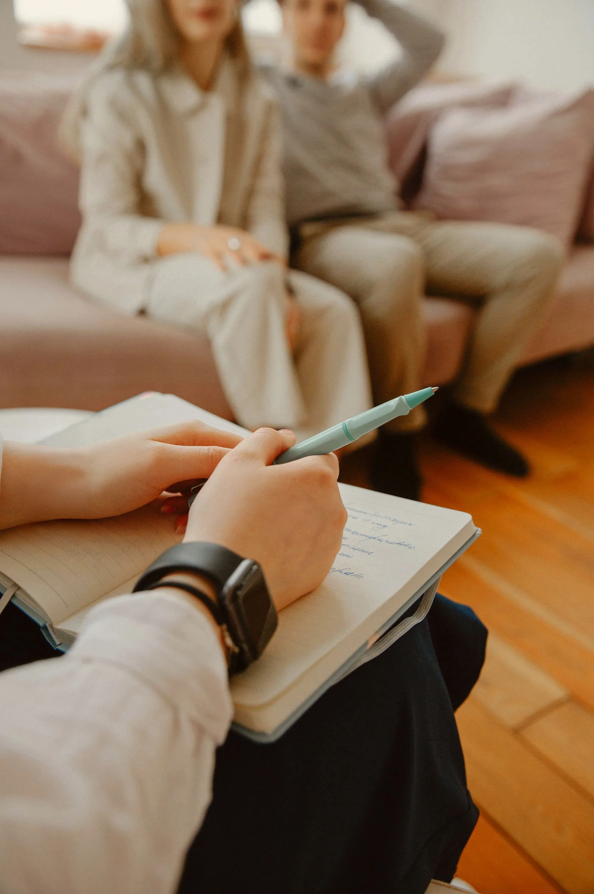 Person taking notes on a notepad with a teal pen, sitting on a chair with a black watch on their wrist, while two people are sitting on a sofa in the background.