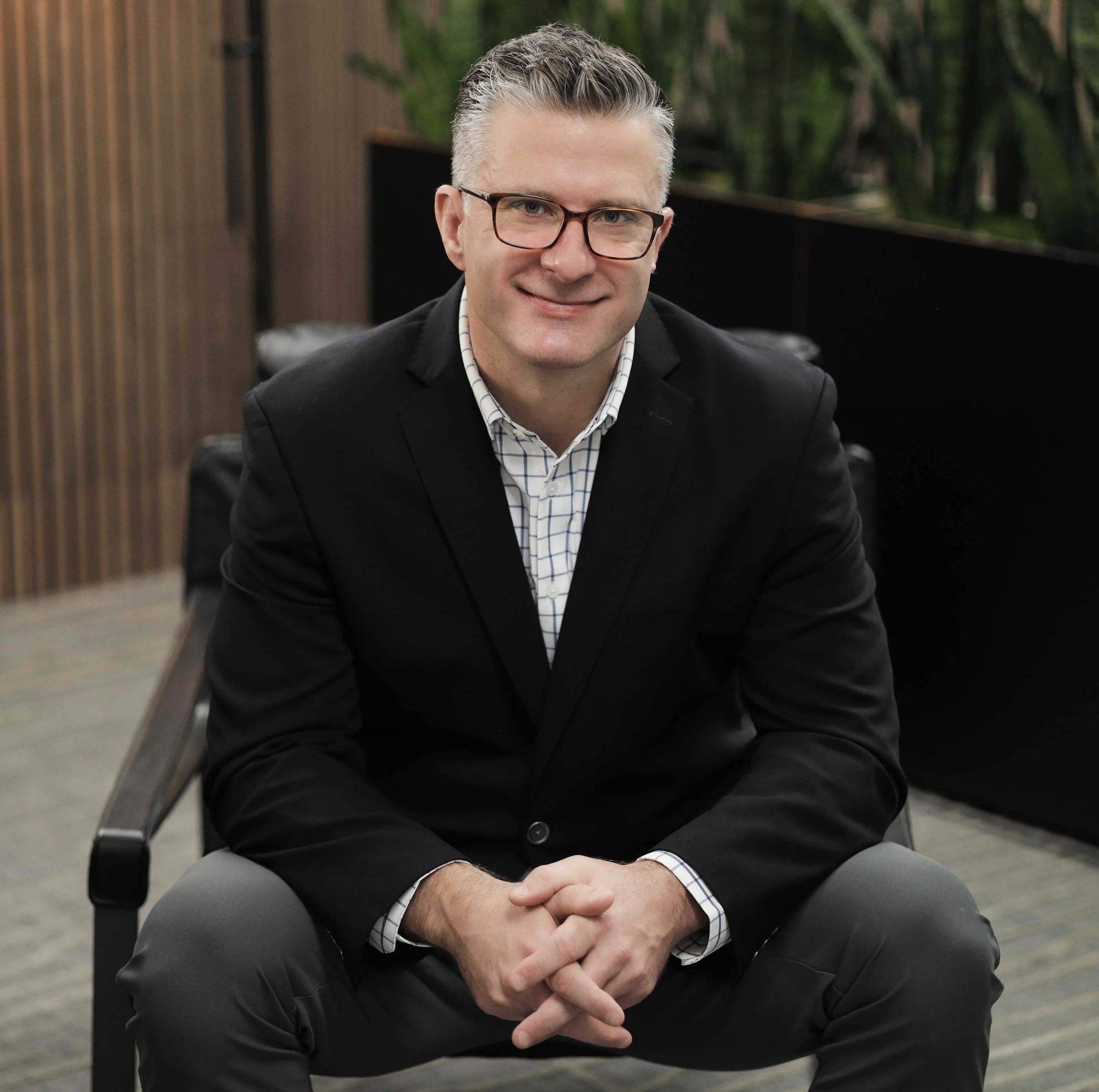 A man with glasses, gray hair, wearing a black blazer and a checkered shirt, sitting on a chair in an office environment, smiling at the camera.