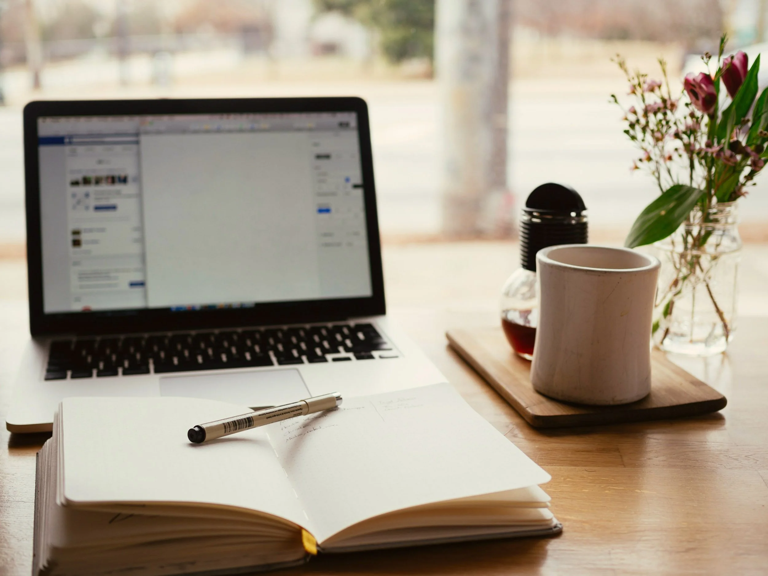 Open notebook with pen, laptop, mug, small glass container with tea, and flowers on a wooden desk near a window.