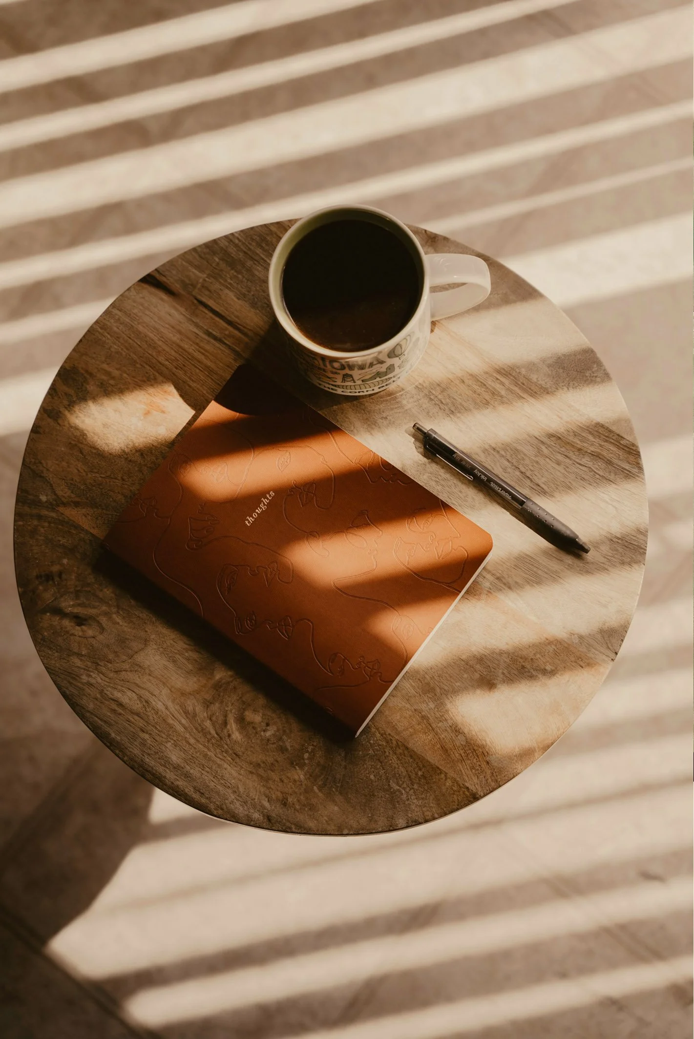 A round wooden table with a mug of coffee, a closed orange notebook labeled 'thoughts,' and a black pen. Sunlight creates striped shadows on the table.