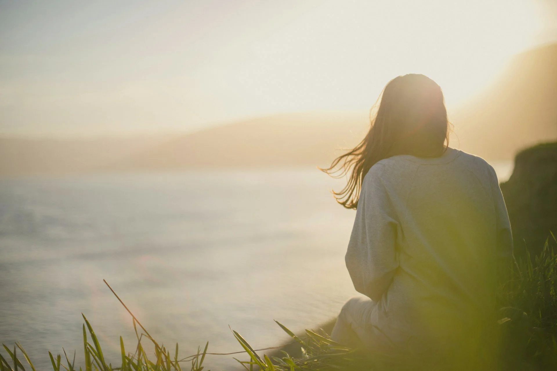 A woman sitting on the grass near water at sunset or sunrise, with her back to the camera and her hair blowing in the breeze.