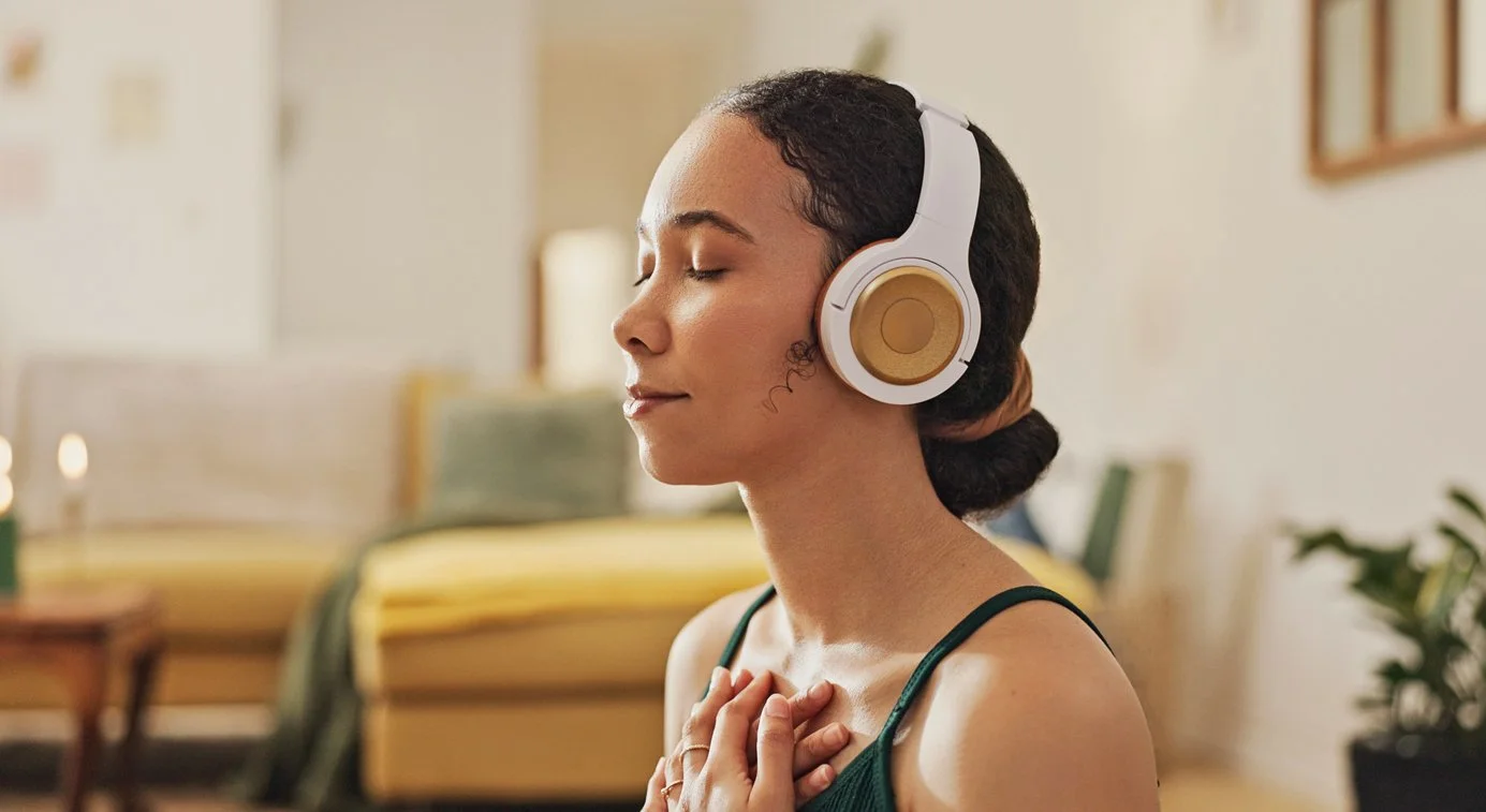 A woman with closed eyes and a peaceful expression wears white and gold over-ear headphones, with her hands placed over her chest, in a cozy indoor setting.