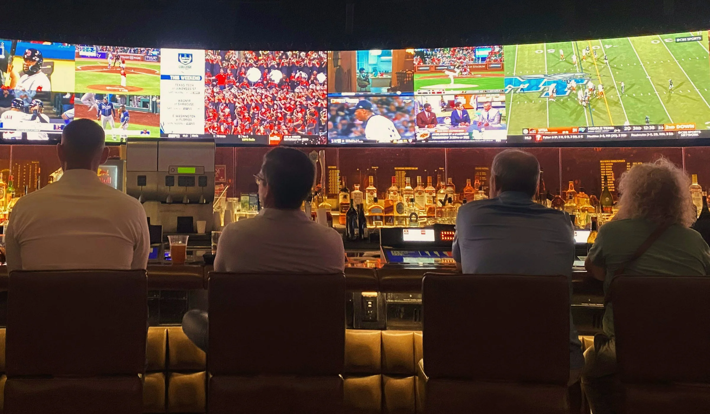 Four people sitting at a bar watching multiple television screens showing various sports events.