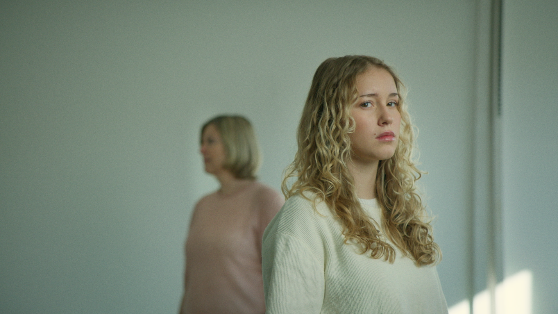 Zwei Frauen stehen vor einem Spiegel, eine im Vordergrund mit lockigen Haaren, die andere im Hintergrund mit kurzem Haar. Die Frauen sind in einem Raum mit neutraler Wandfarbe.