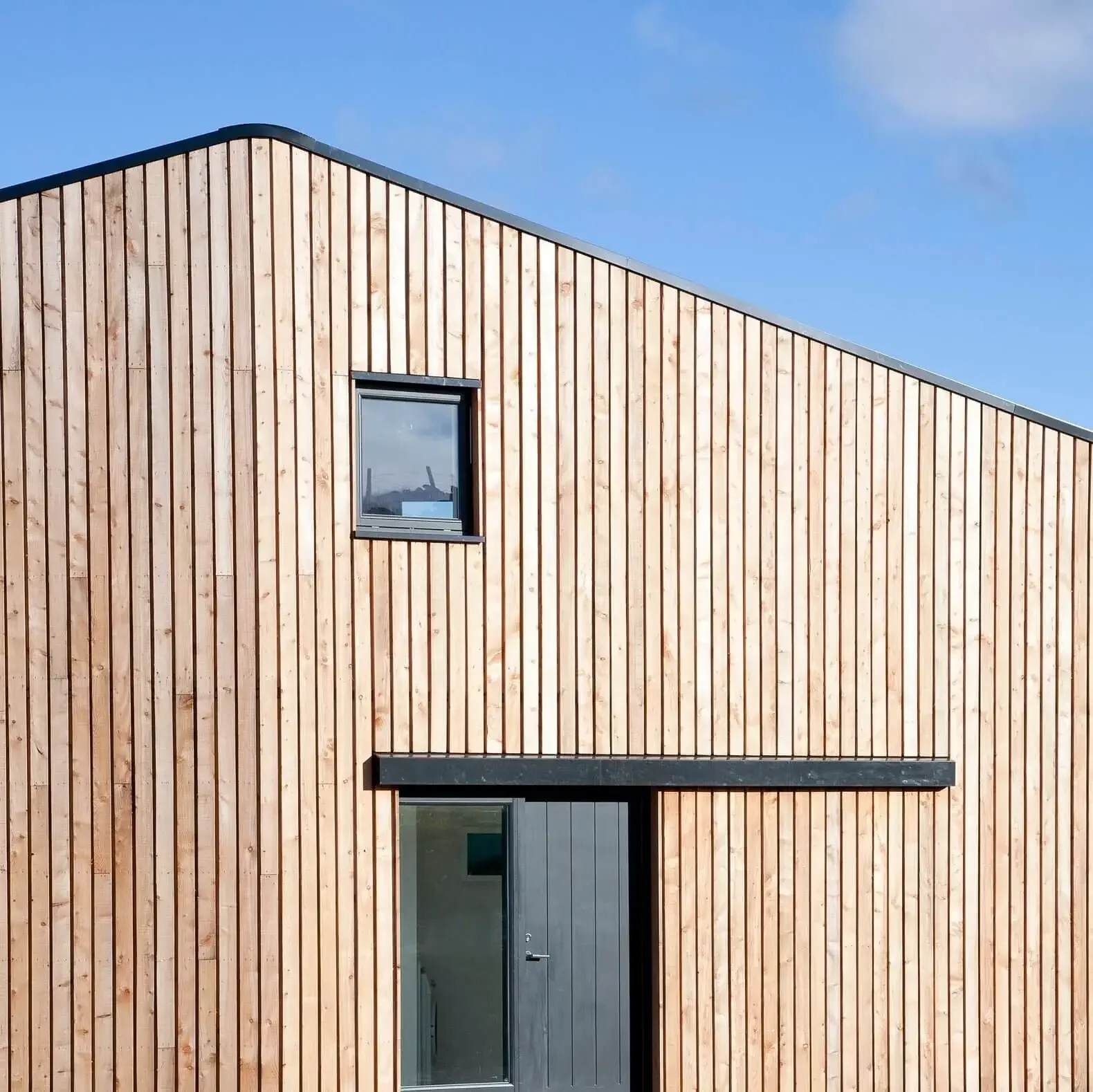 Modern building with vertical wooden slats, two windows, and a black door set against a blue sky.