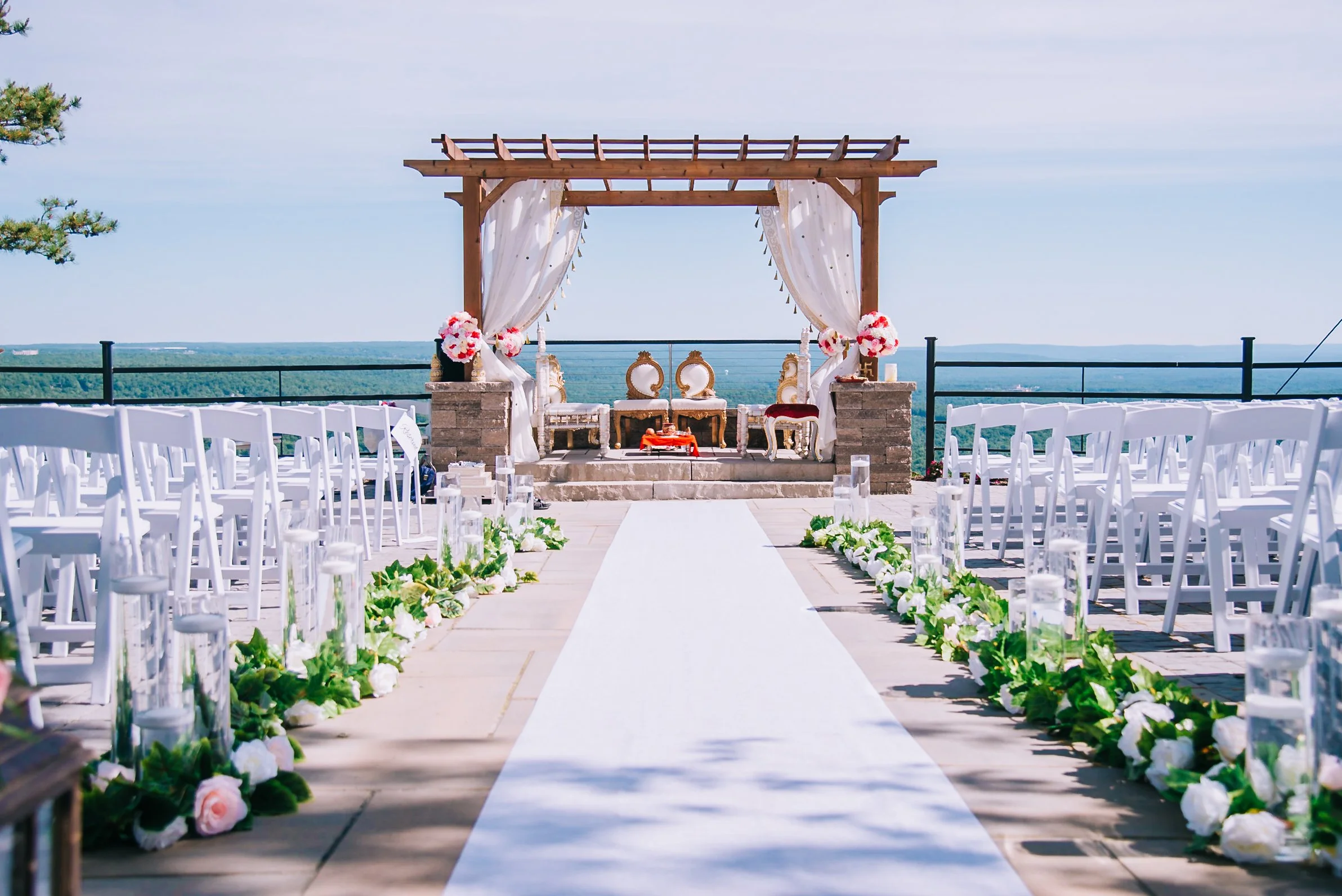 Outdoor wedding ceremony setup on a deck overlooking the ocean, with white chairs arranged on either side of a white aisle runner, floral arrangements, and a wooden arch with white curtains.