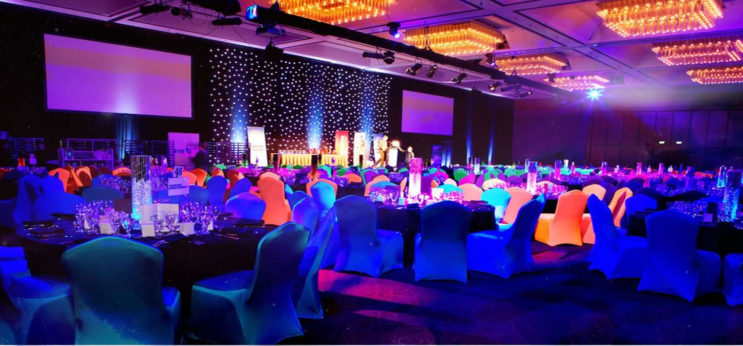 A hotel ballroom decorated for a formal event with round tables covered in black tablecloths and colorful chairs, a stage with a backdrop of black and white stars, and large projection screens. The tables are set with glassware, candles, and centerpieces, and the room is illuminated with blue and purple lighting.