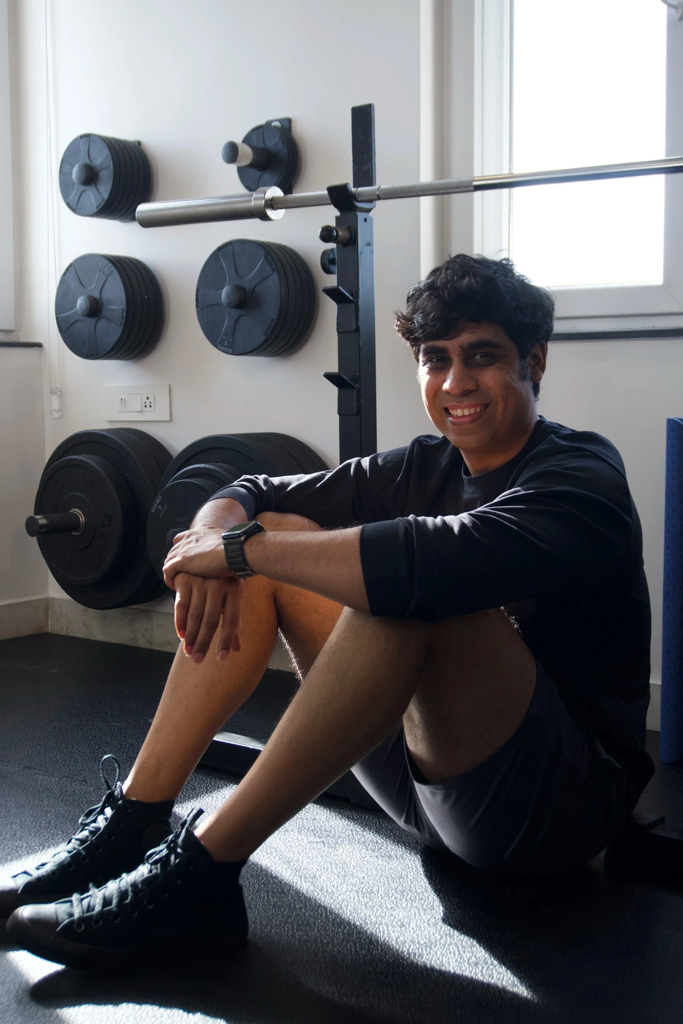 Personal trainer Chirag Chordia smiling while sitting on the rubber gym flooring next to a weight rack in Chennai.