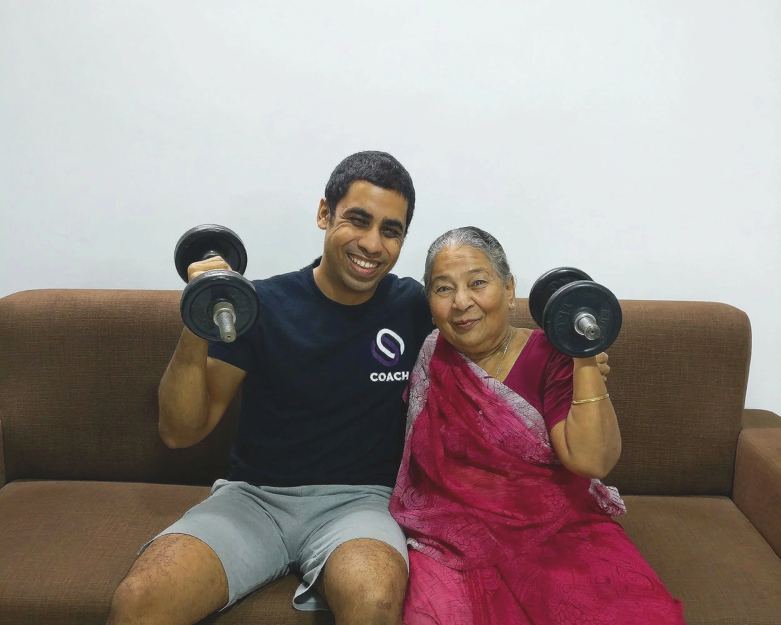 Chirag Chordia holding dumbbells on a sofa next to his grandmother, Kiran Bai, in a warm home setting.