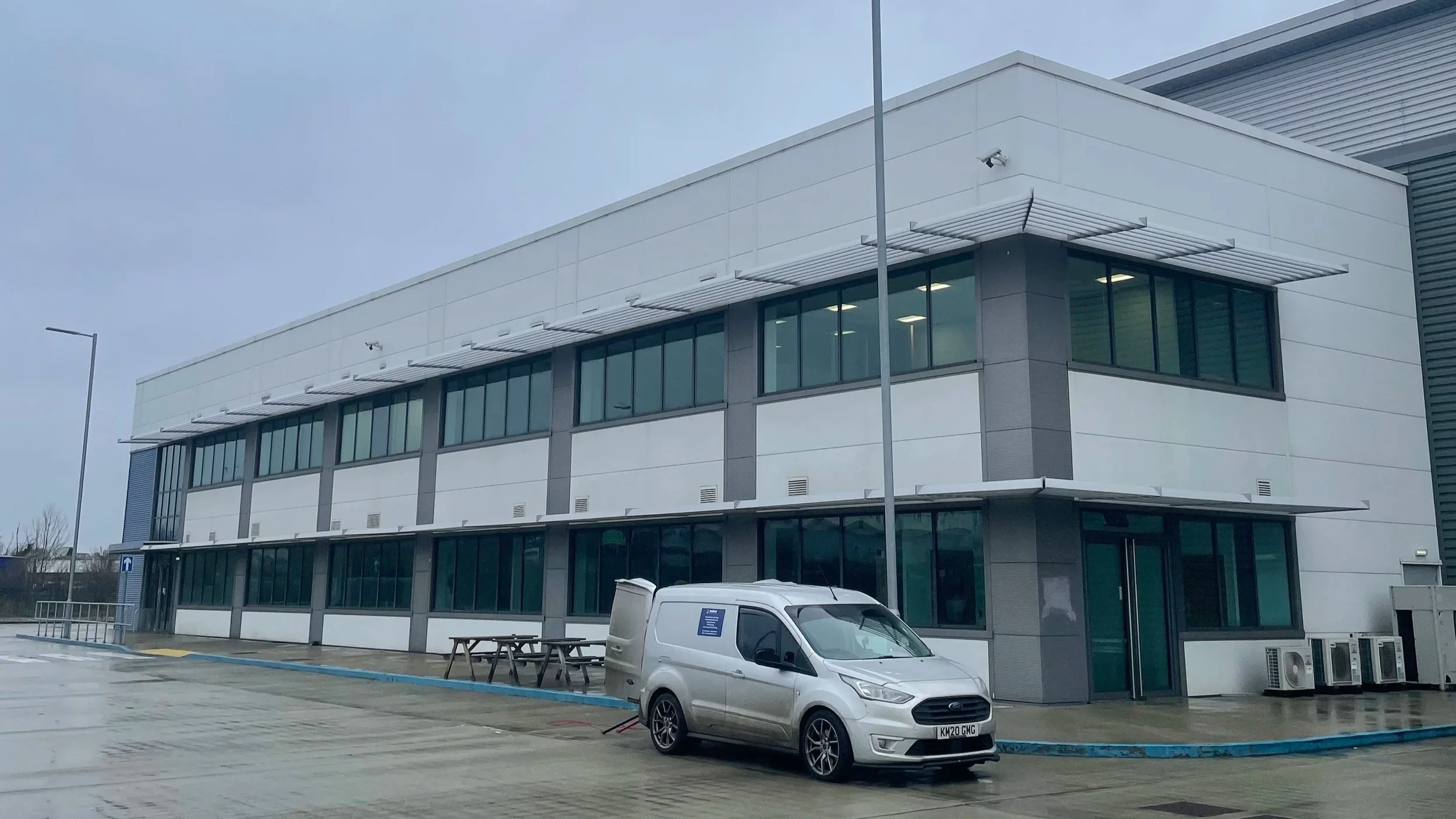 A modern, multi-story office building with large tinted windows, metal awnings, and a parking lot in front, featuring a silver van parked near picnic tables on an overcast day.