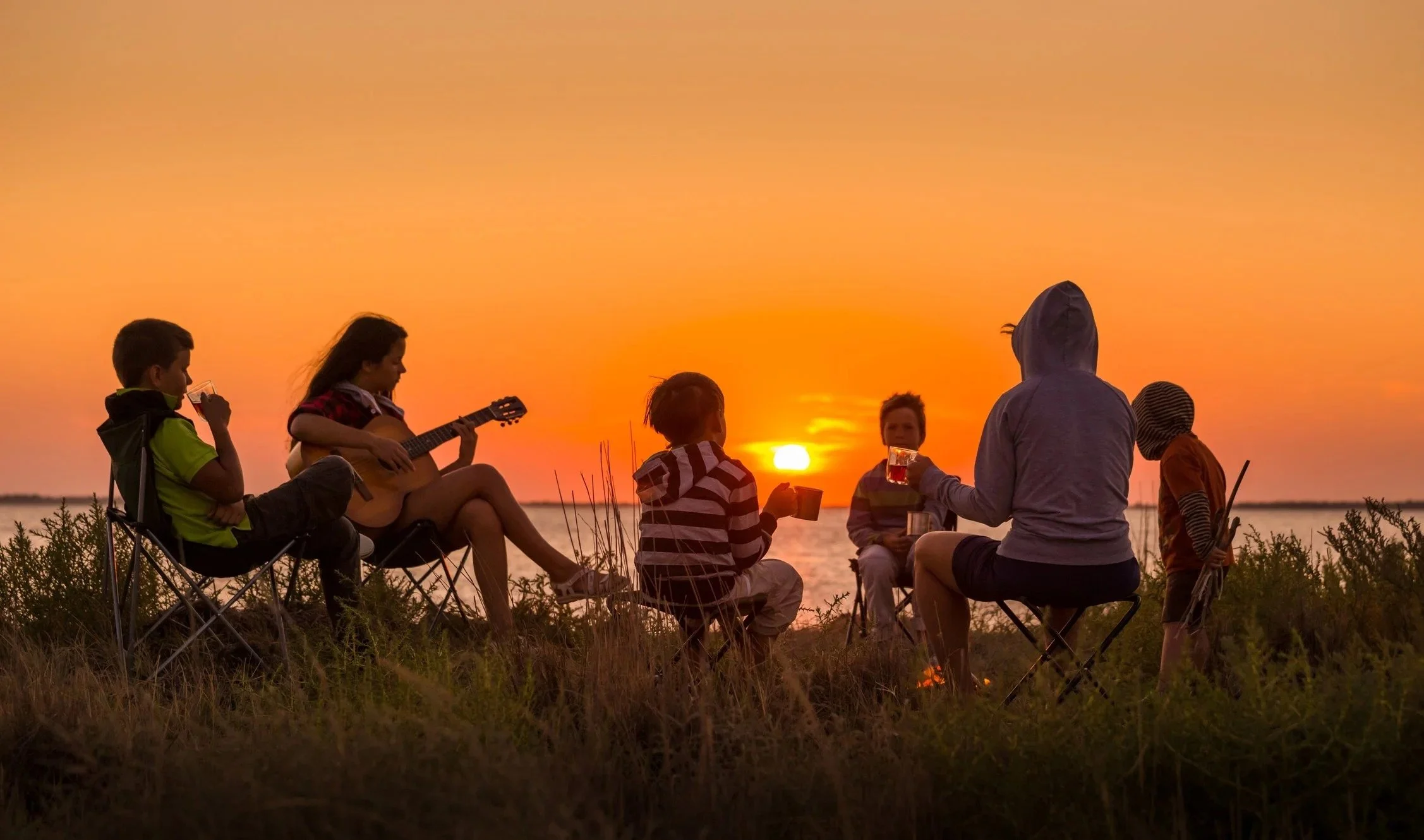A step family sitting on camping chairs and small stools by a body of water during sunset, some playing guitar and others holding drinks or cups, with vegetation in the foreground.