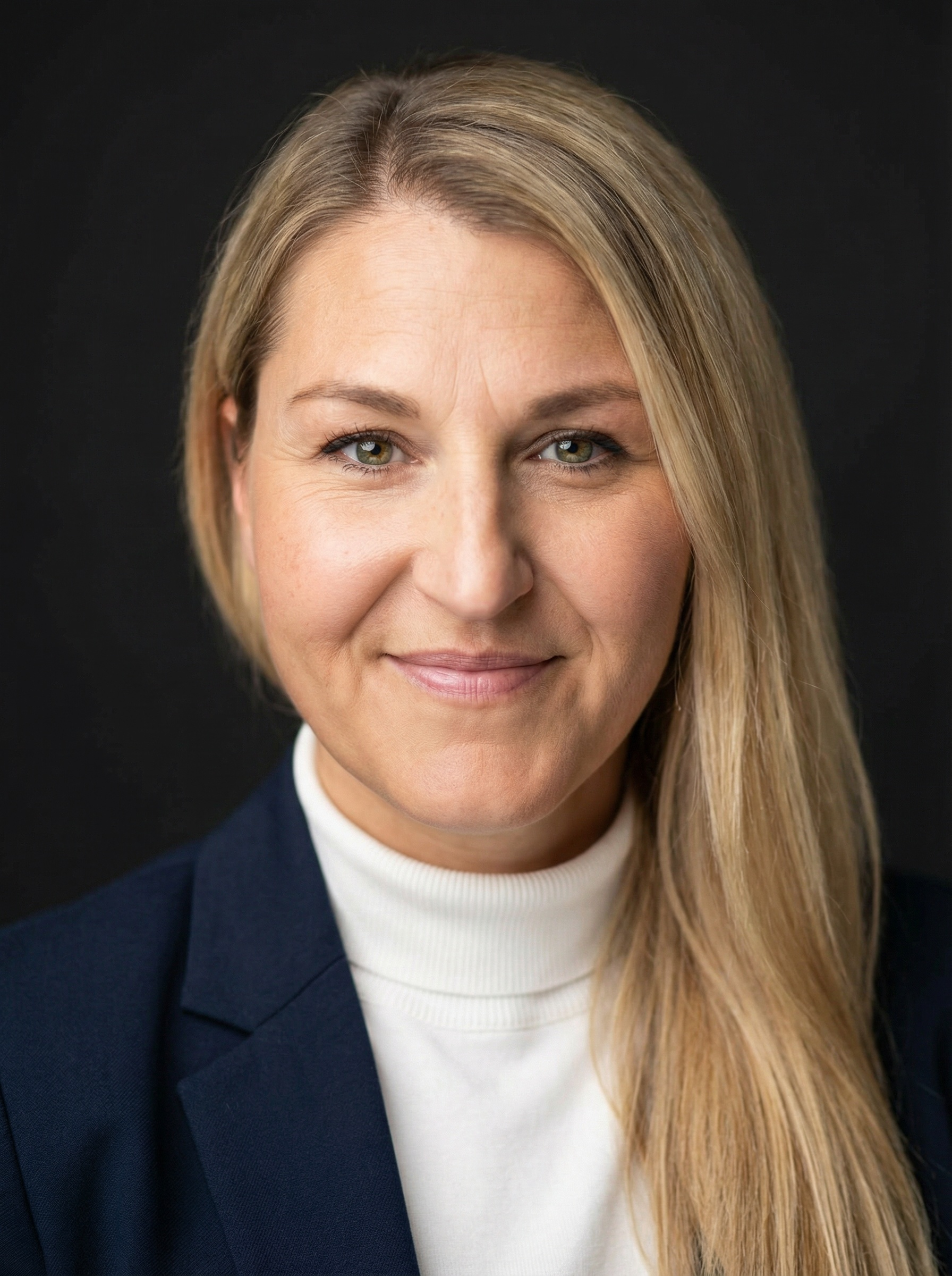 A professional headshot of a woman with blonde hair, wearing a navy blazer over a white turtleneck, smiling subtly against a dark background.