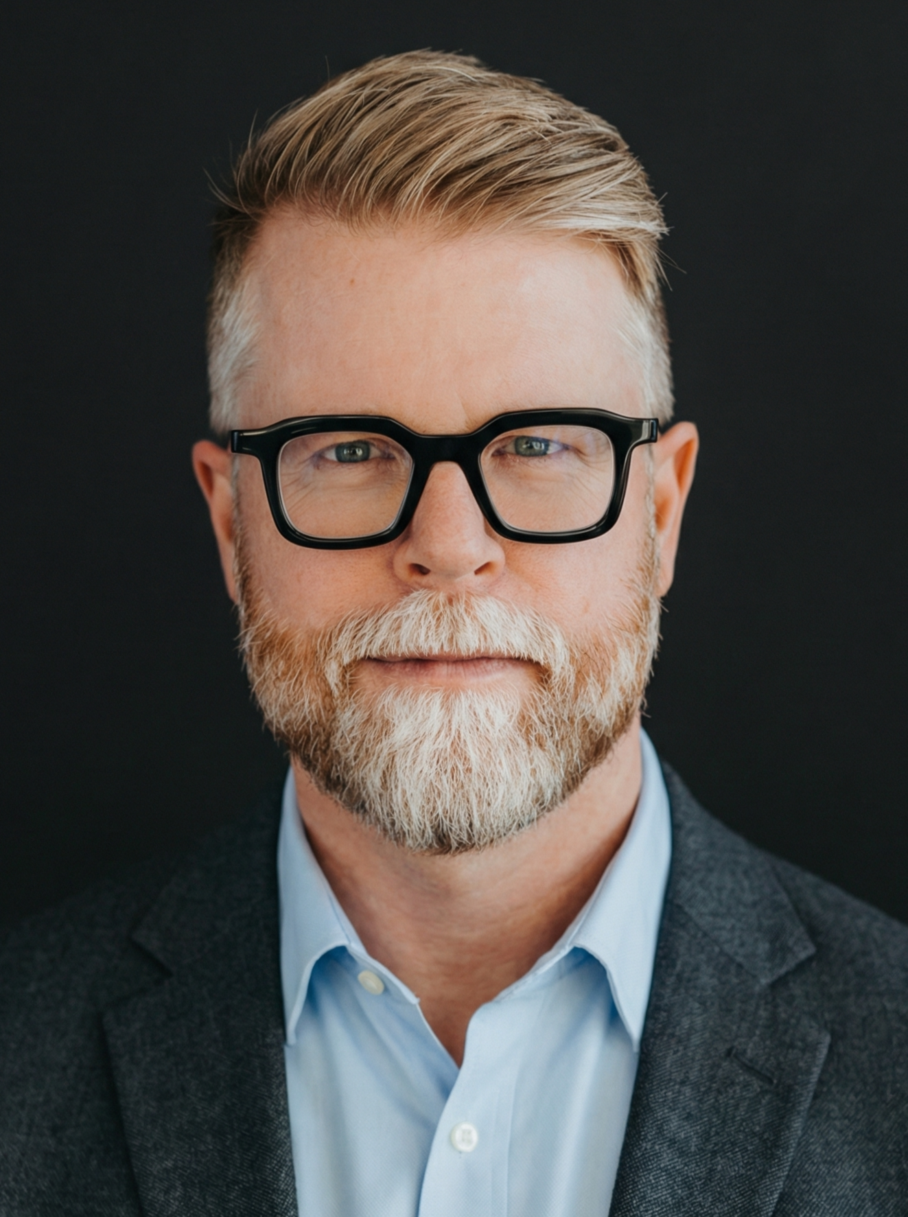 Portrait of a mature man with glasses, a beard, and blonde hair, wearing a blue shirt and a dark blazer, against a dark background.