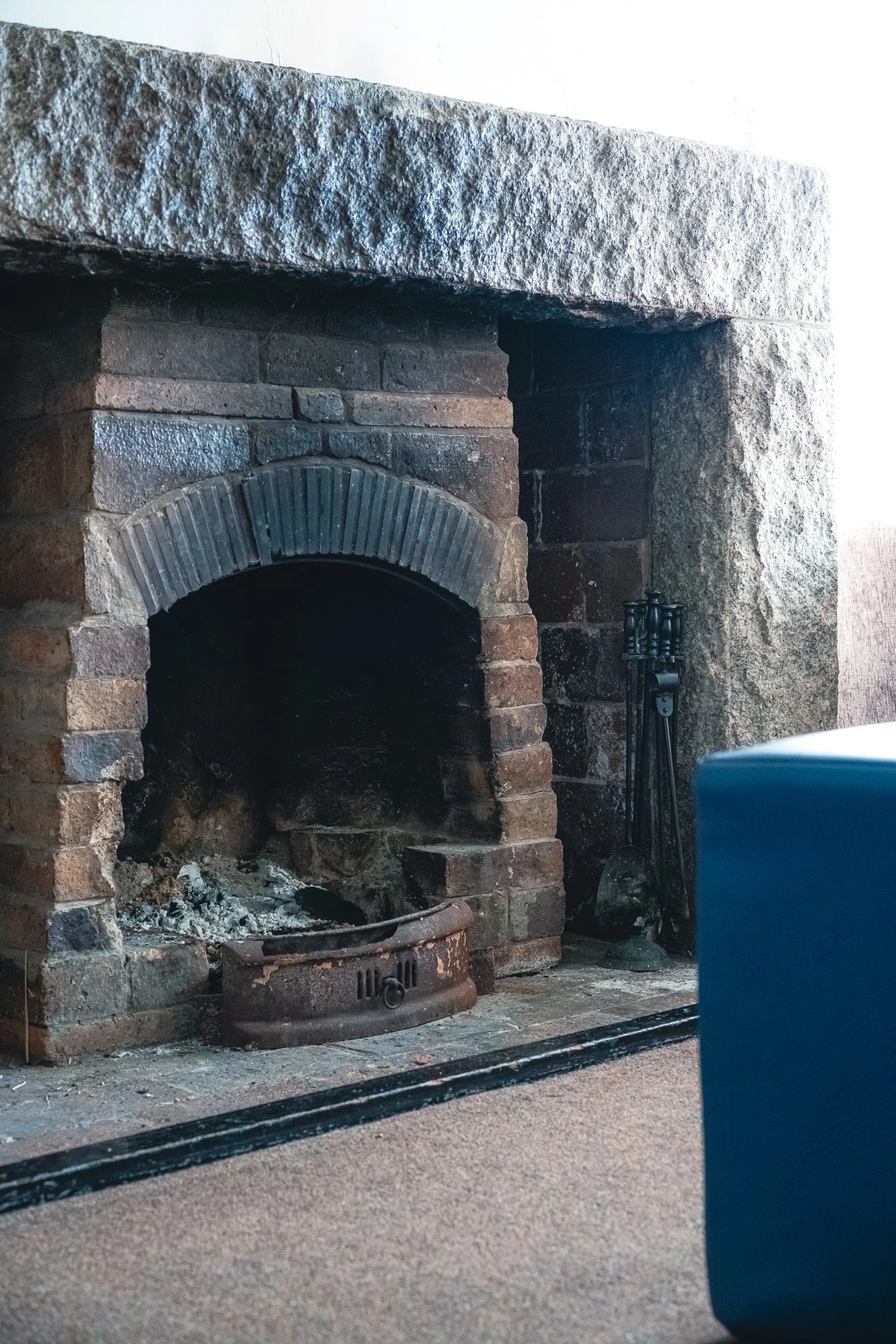 A rustic stone fireplace with a brick arch and a metal fire grate, set against a stone wall. Fireplace tools are on the right side.