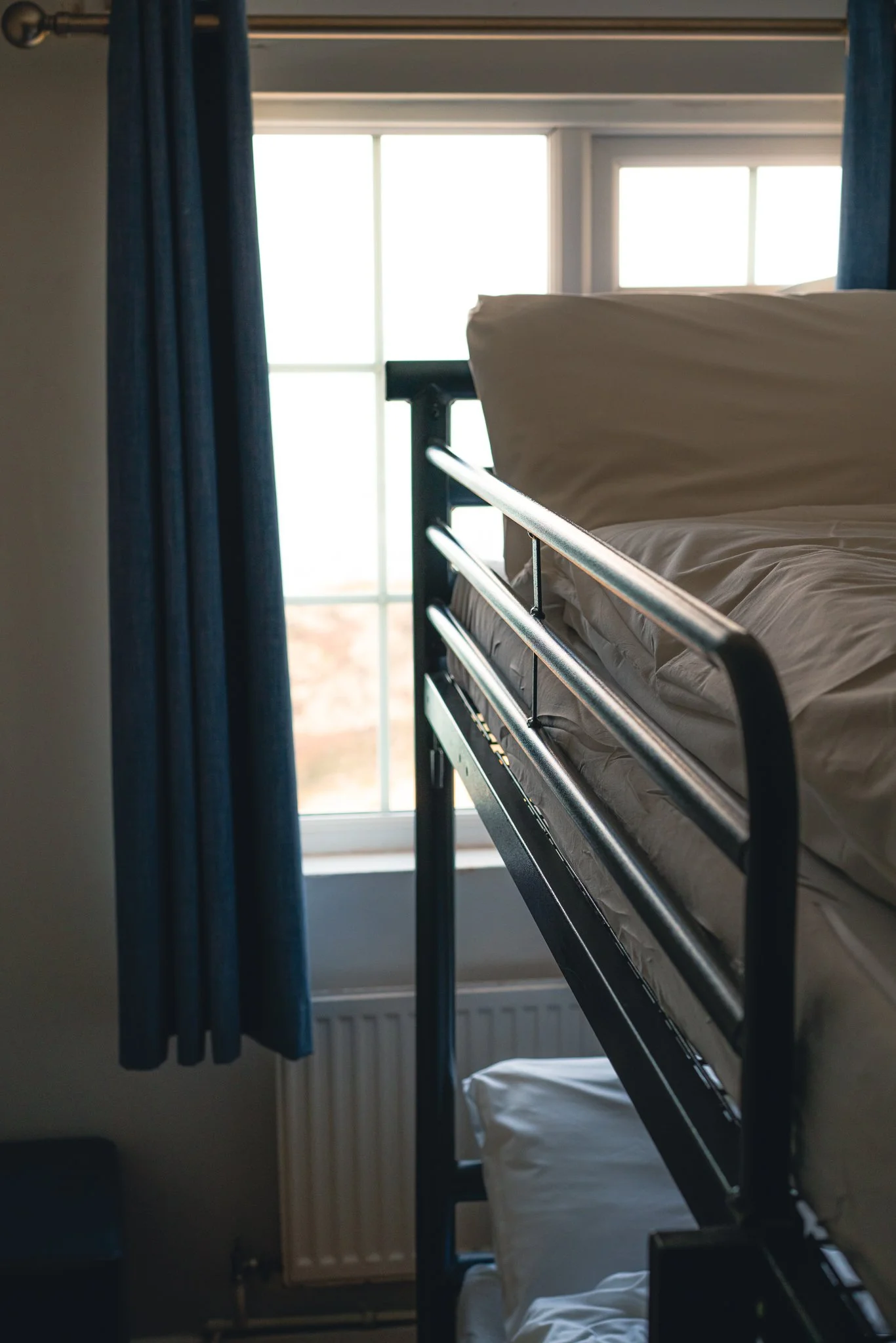 An upstairs bedroom with a bunk bed near a window with white framing and blue curtains. The bed has white sheets, and there is a radiator beneath the window.