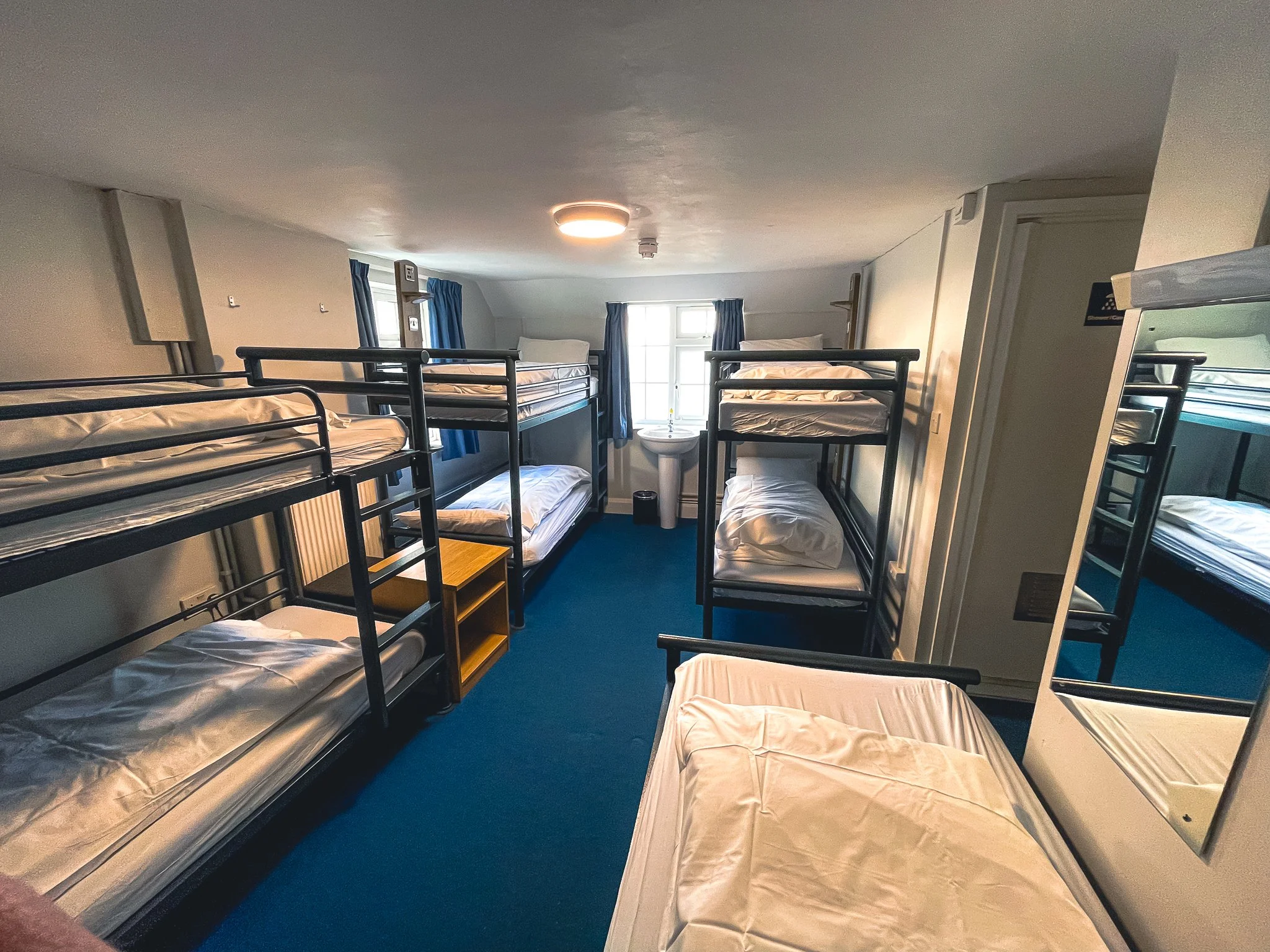 Multiple bunk beds with charging facilities, white mattresses and bedding. The room has blue carpet, a window with blue curtains, and a ceiling light.