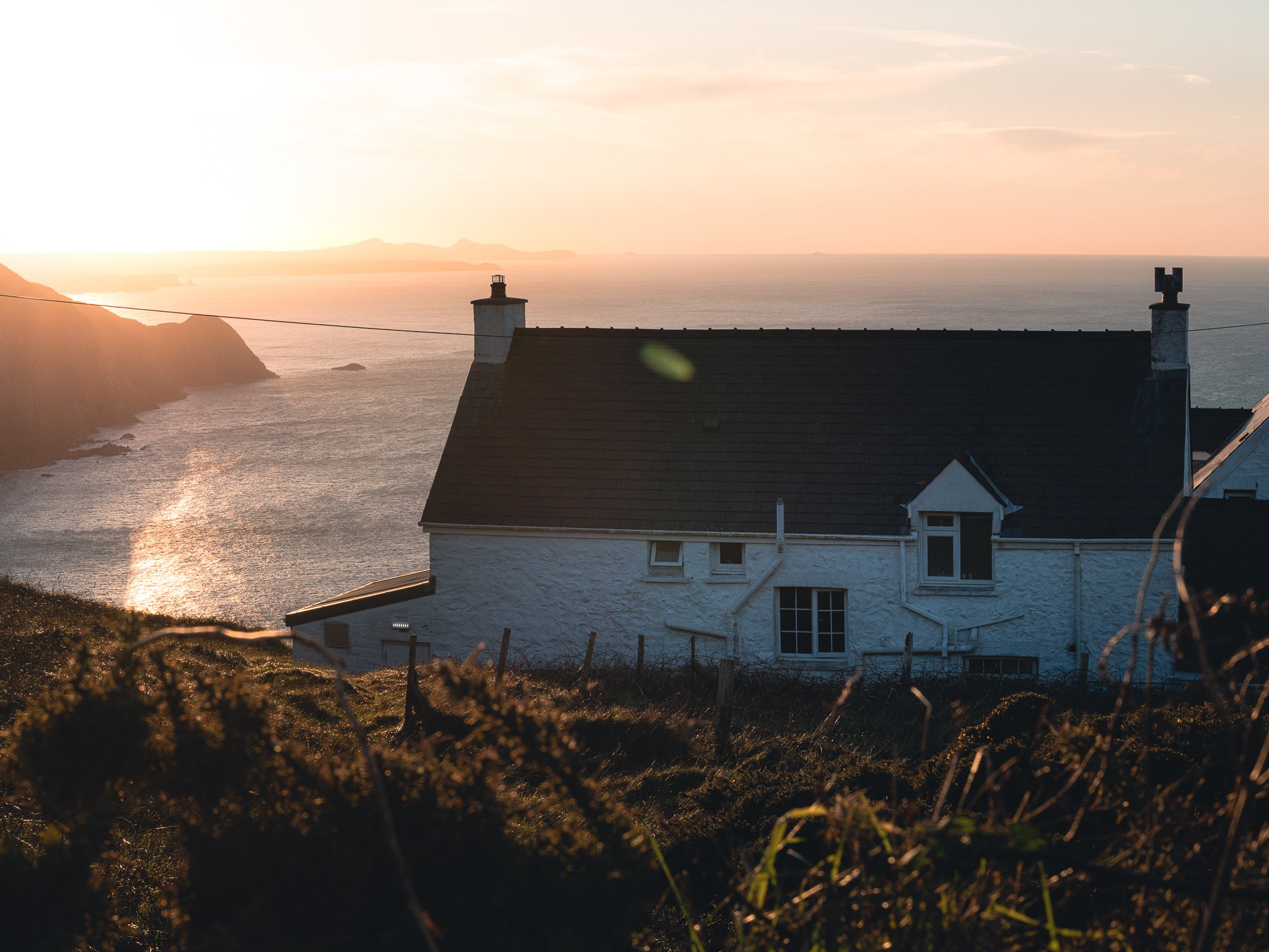 A house with a dark roof and white walls situated on a grassy hillside overlooking the ocean at sunset, with cliffs on the coastline in the background.