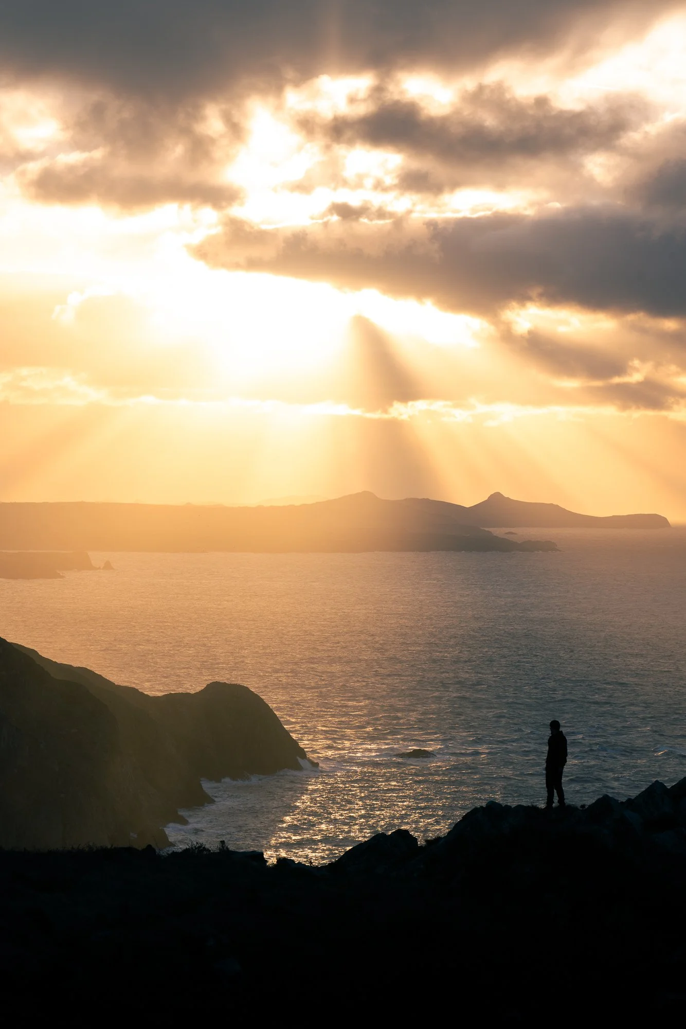 A sunset over the ocean with rays of sunlight breaking through the clouds. A person is standing on rocks near the water, looking out at the scene with mountainous land in the background.