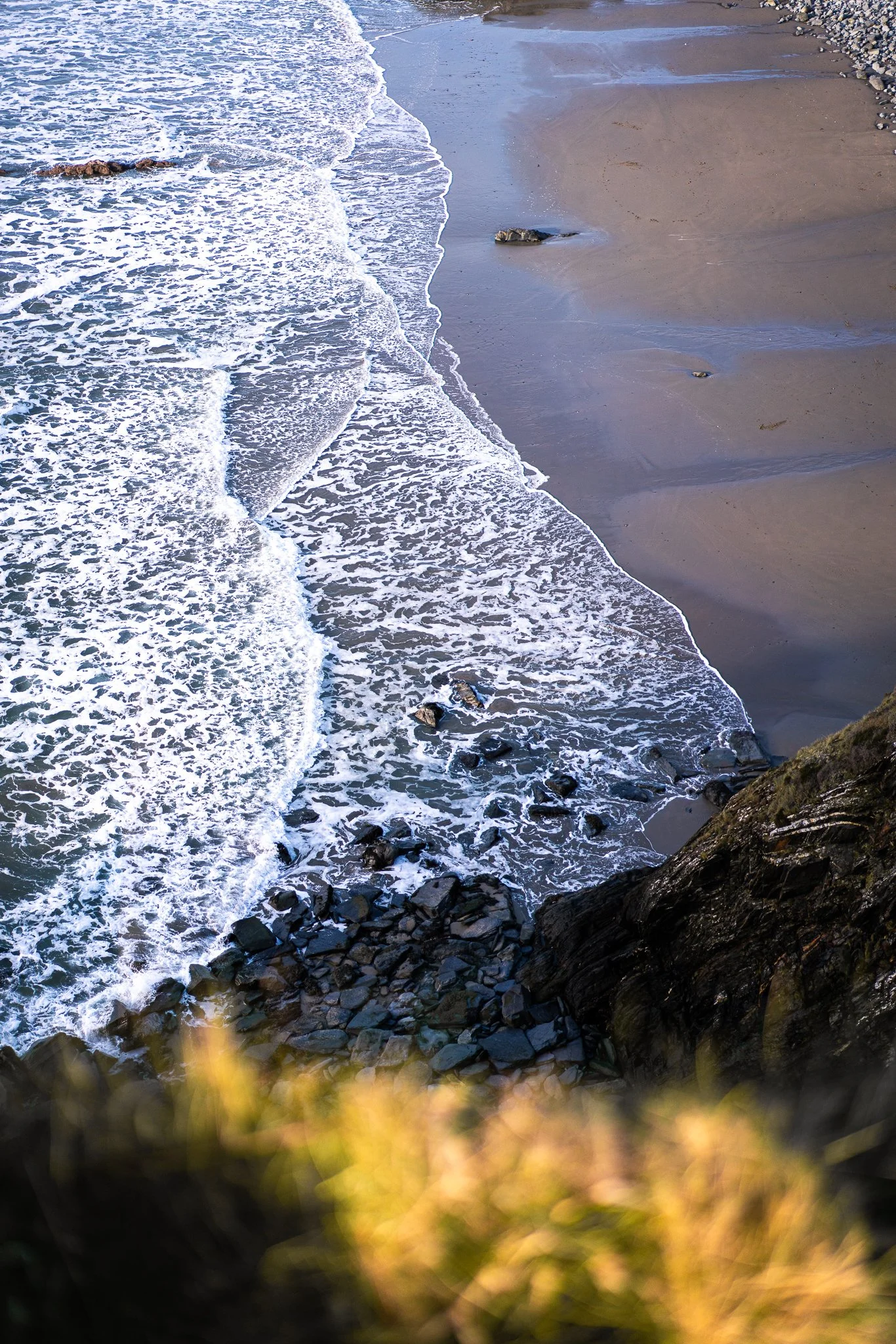 View of a sandy beach with rocks and ocean waves crashing along the shoreline, taken from a hillside.