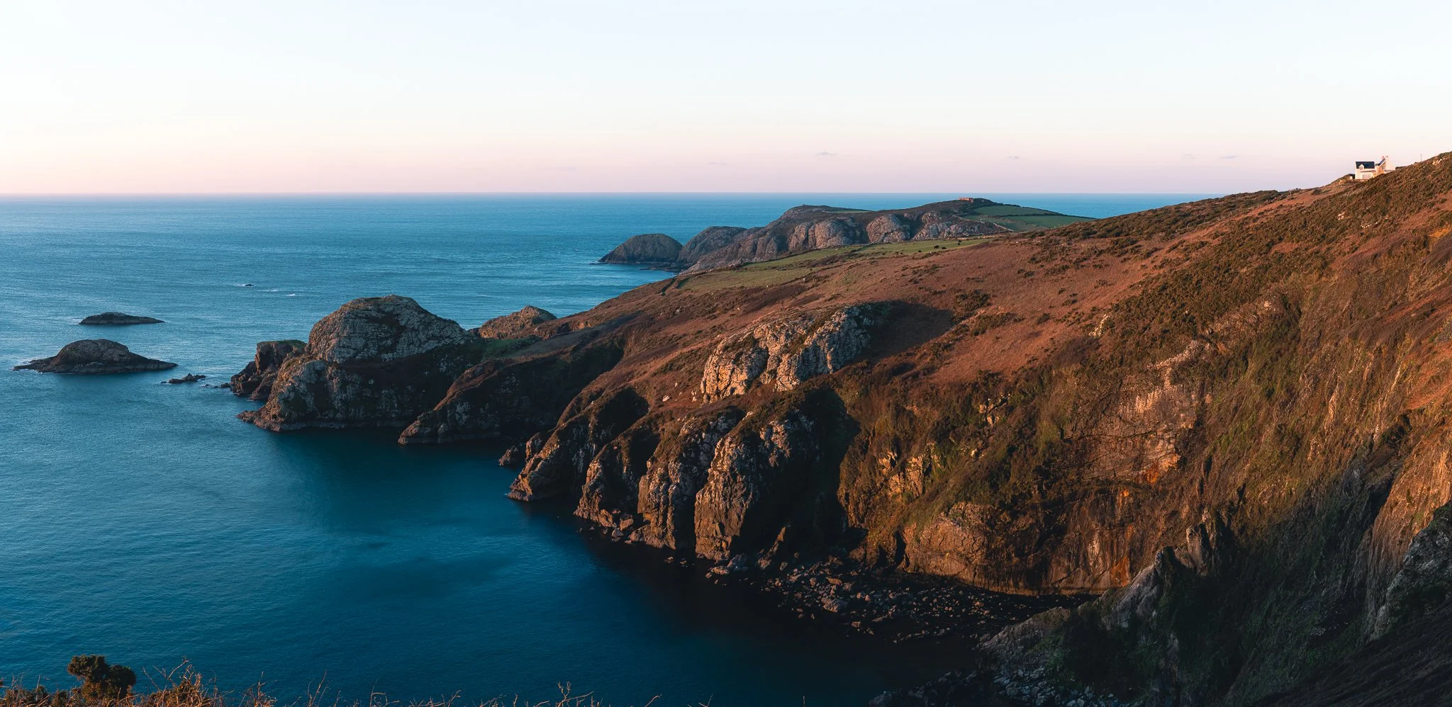 Scenic view of rugged coastal cliffs along the ocean during sunset with a house on the top right.