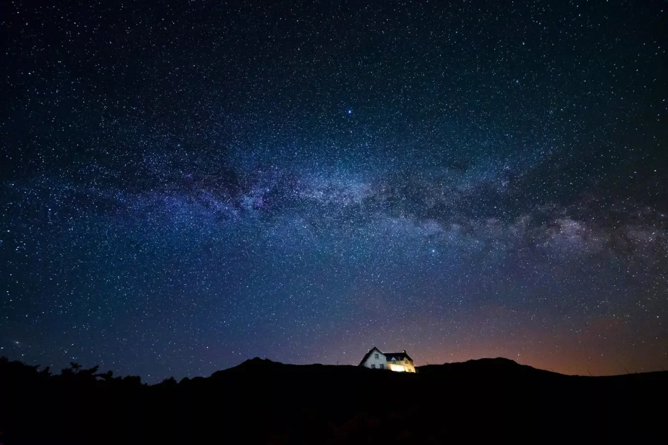 Night sky filled with stars and the Milky Way galaxy, with a house illuminated in the dark landscape below.