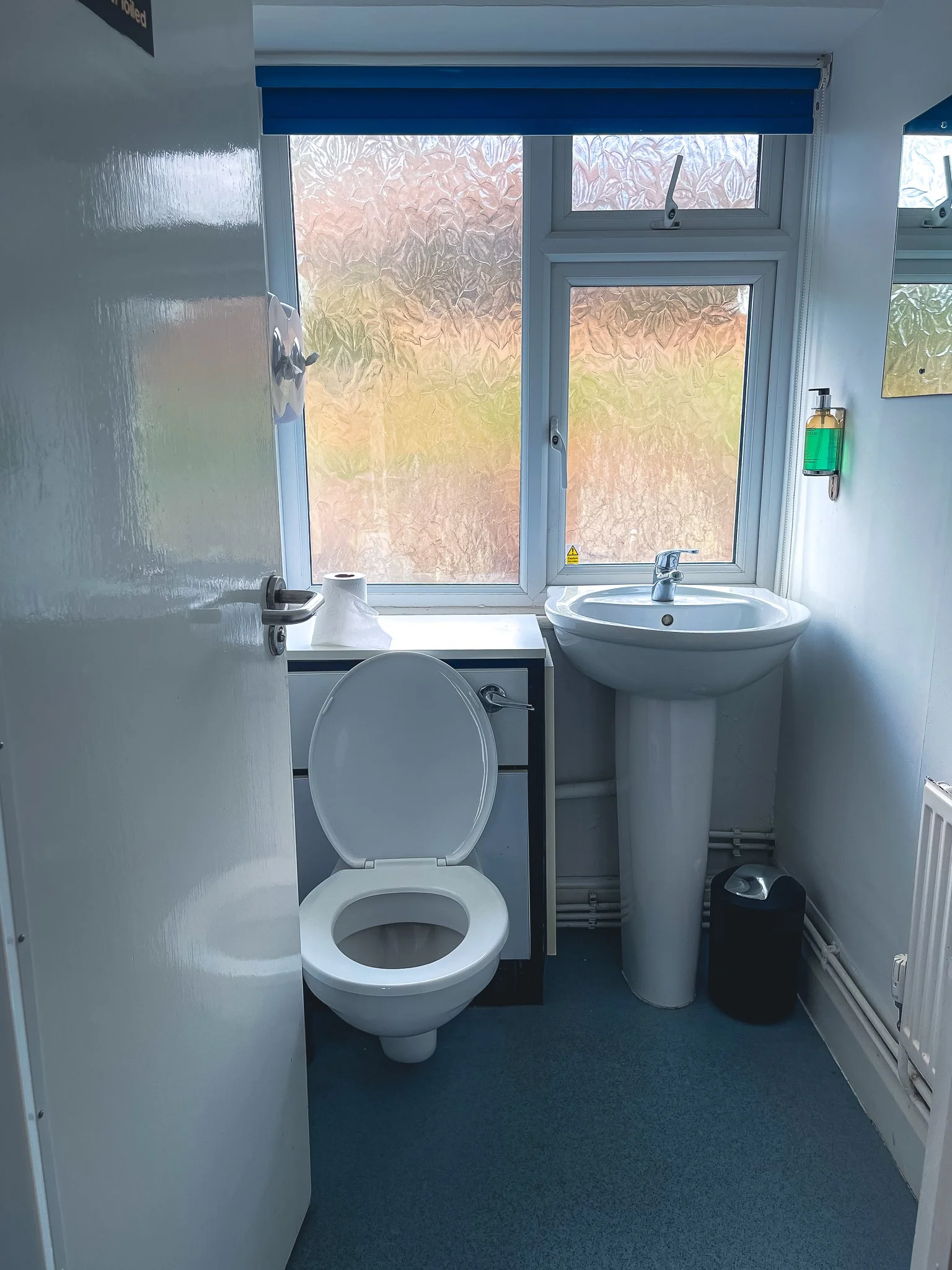 A small bathroom with a toilet, a sink with a faucet, a mirror, a shelf with a bottle of hand soap, a trash bin, and a window with frosted glass. There are paper towels on top of a cabinet and a partially opened door.