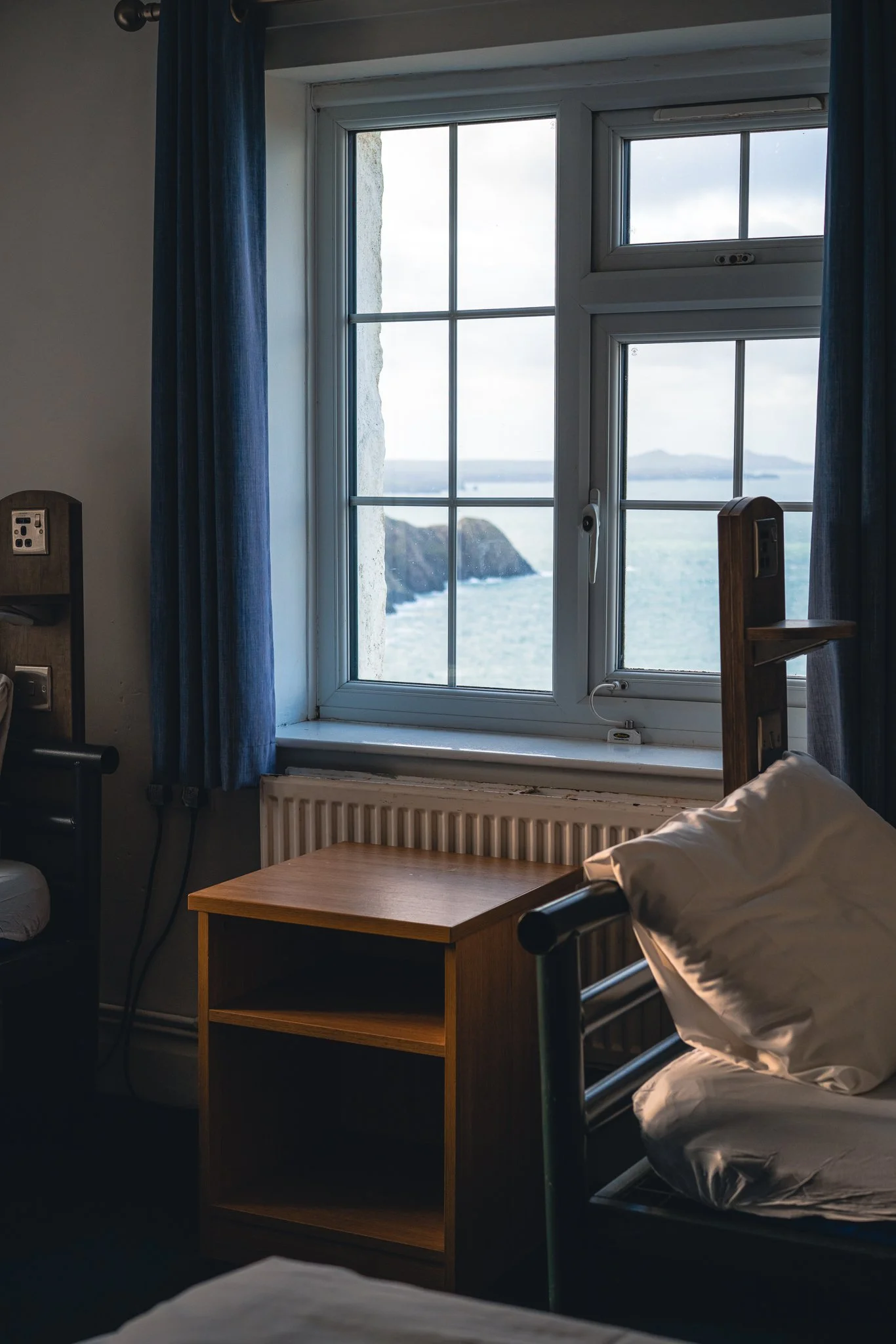 Bedroom with a window overlooking a coastline and islands, a bed with white sheets and pillow, a wooden bedside table, and blue curtains.