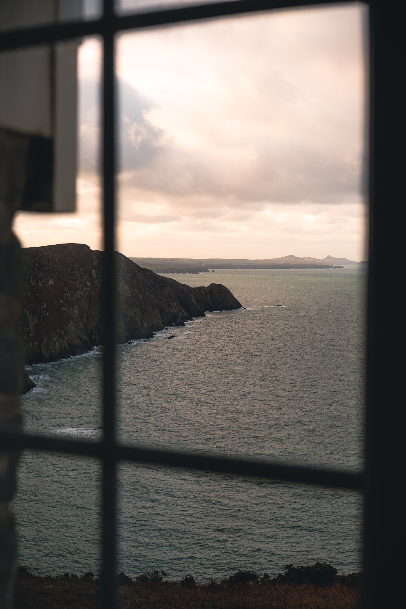 View of a rocky coastline with cliffs and the ocean, captured through a window, under a semi-cloudy sky with sunlight breaking through.
