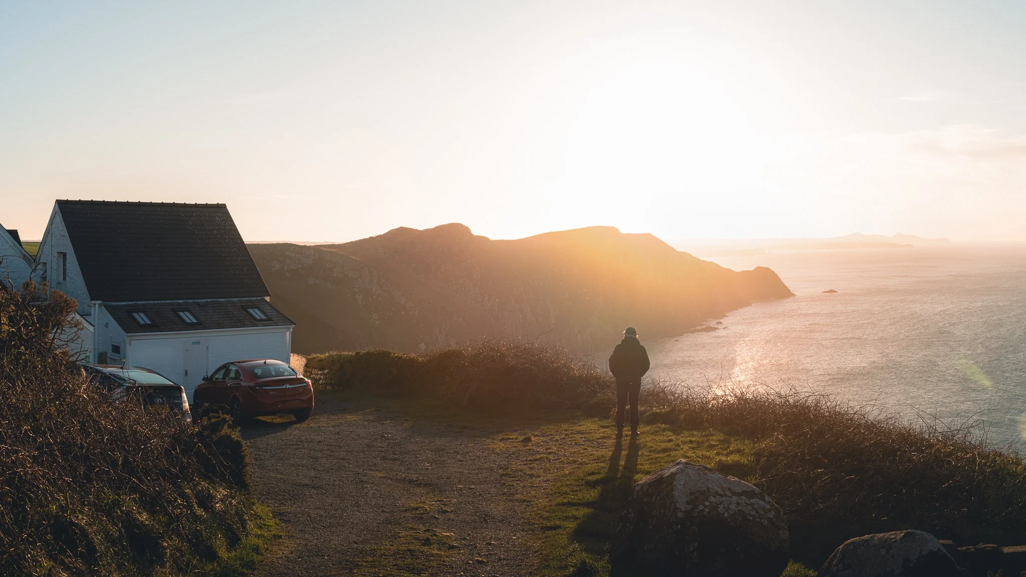 A person standing on a grassy path overlooking the ocean at sunset, with mountains in the background, a house and parked cars nearby.