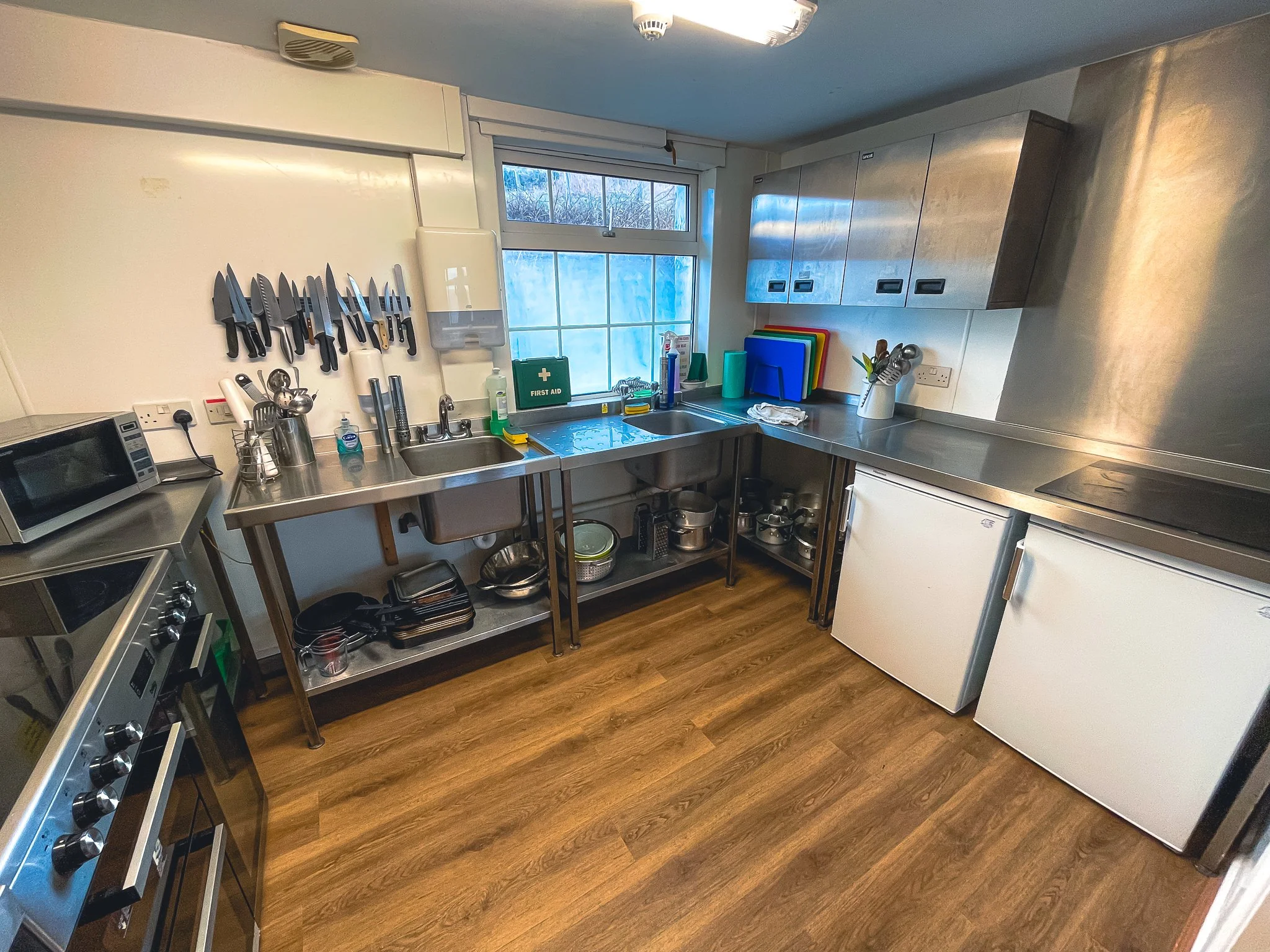 Kitchen with stainless steel countertops, drawers, cabinets, and various kitchen utensils and appliances. A window above the sink shows an outdoor scene. There are knives on a magnetic strip on the wall, cutting boards, and containers on the counter.