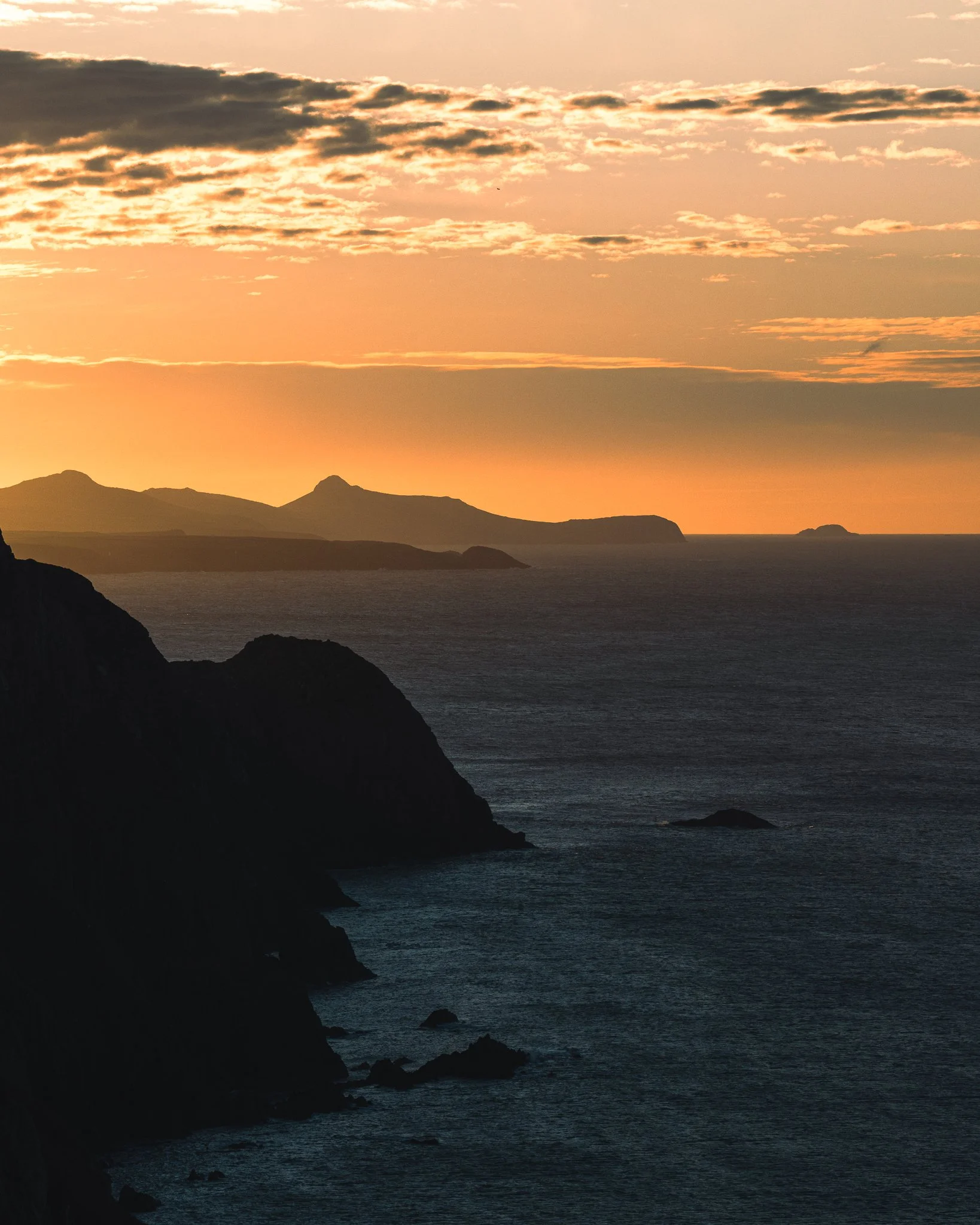 Sunset over a rocky coastline with mountains in the distance and clouds in the sky.