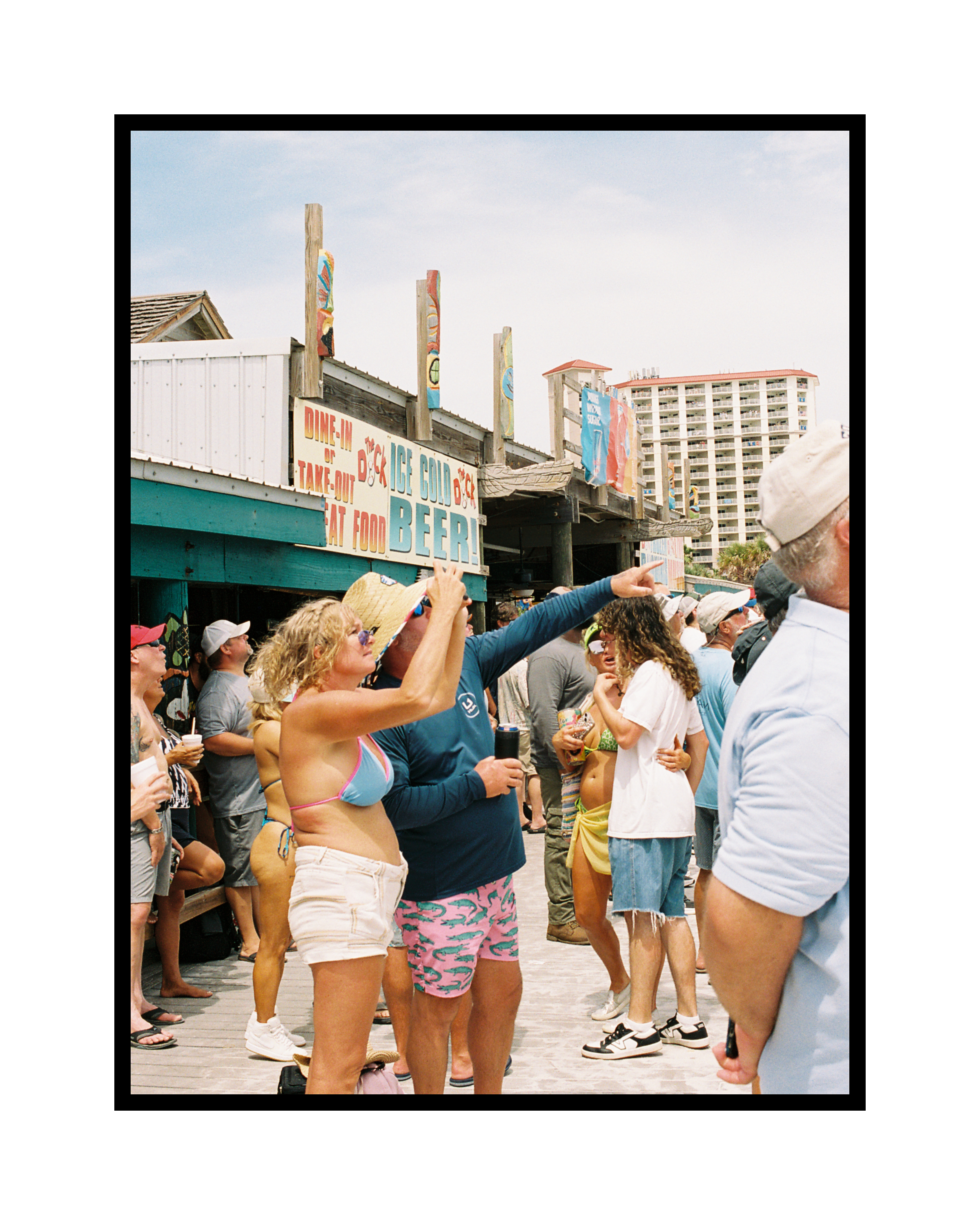 Group of people gathered outdoors near a beach bar, some wearing summer clothes and hats, with a sign advertising food and beer in the background.