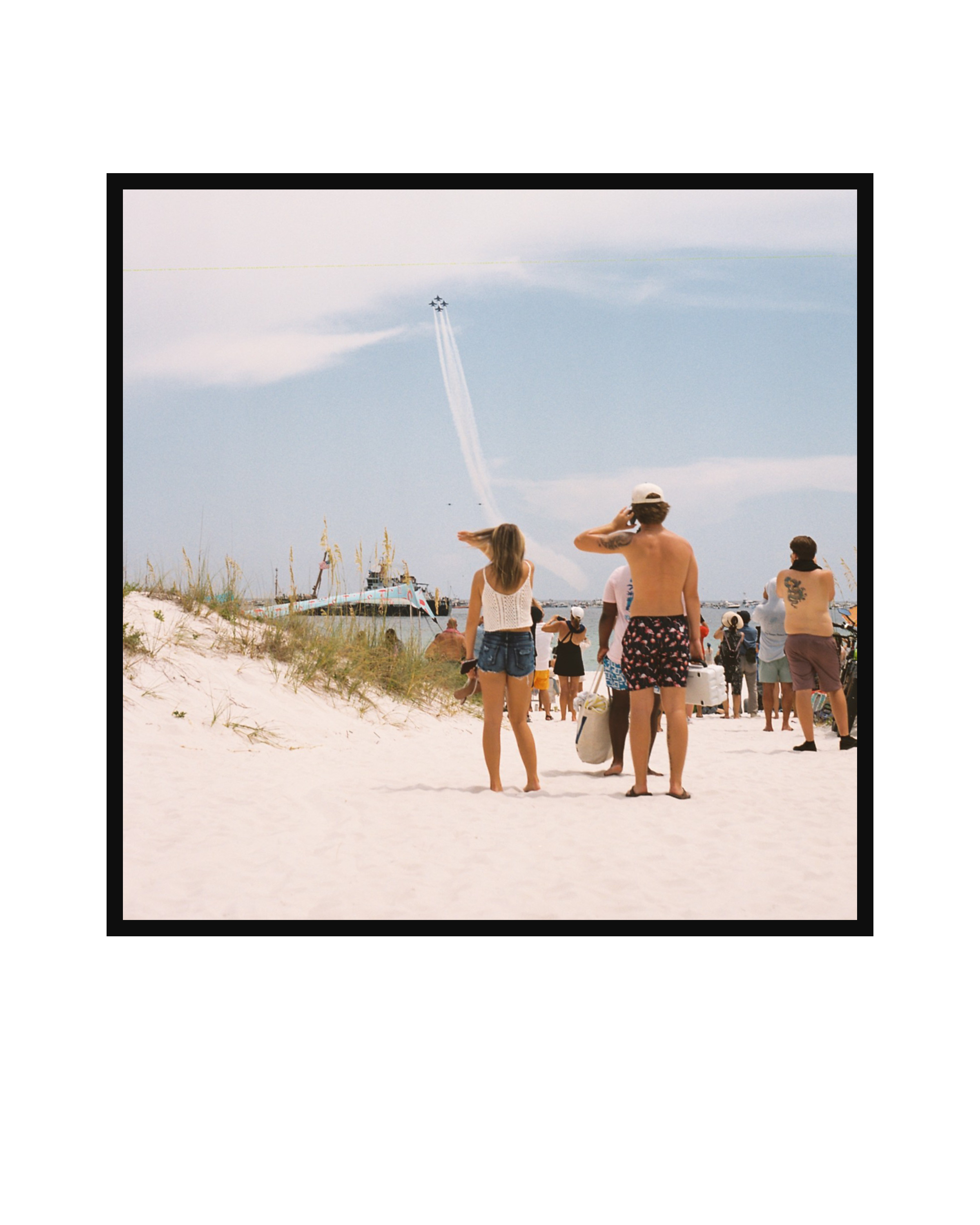 People on a sandy beach watching an air show with the Blue Angels flying in formation and leaving trails in the sky.