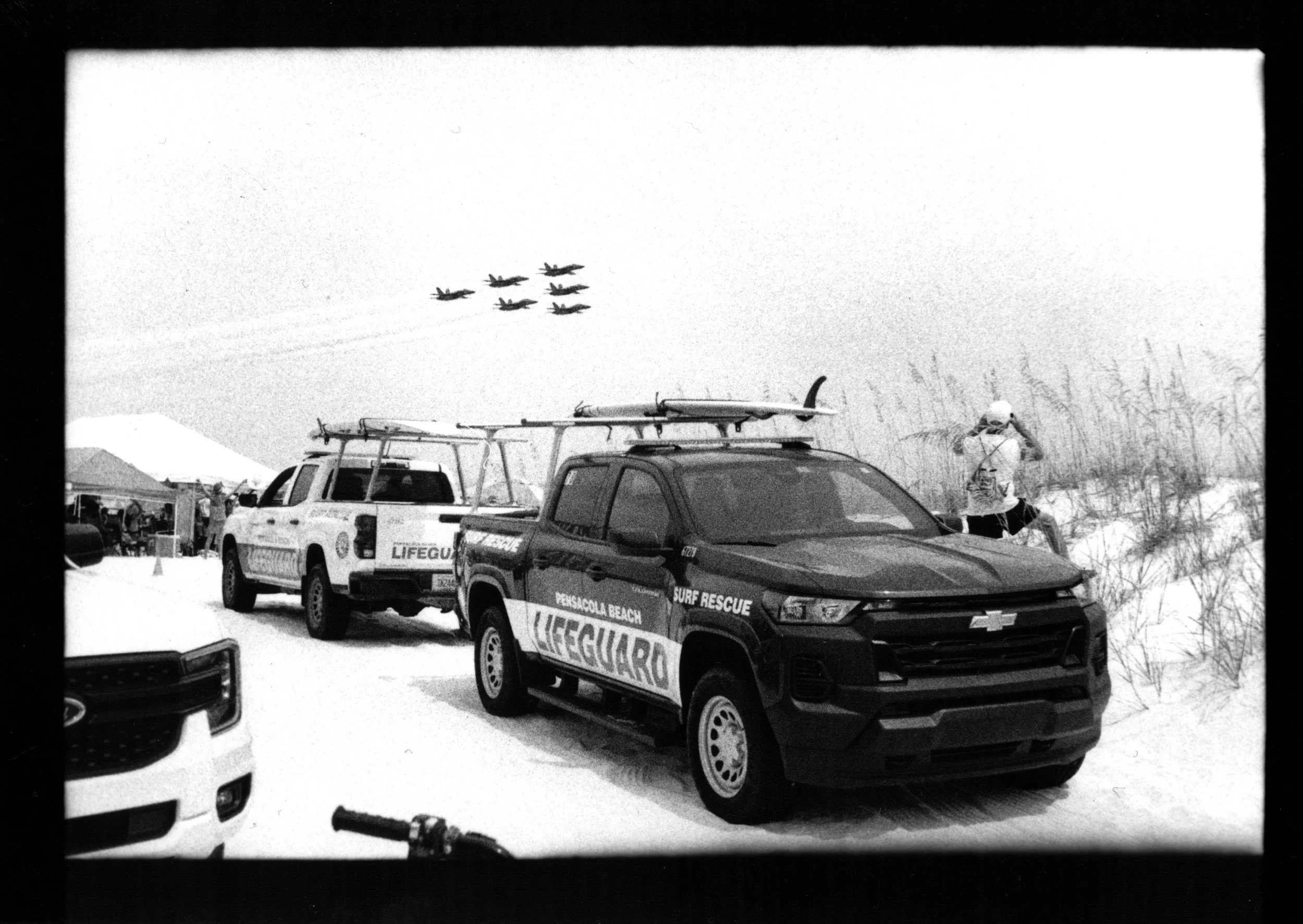 Several airplanes flying in formation over a beach, with rescue vehicles parked on the sand. One vehicle is marked 'Pensacola Beach Lifeguard' and another 'Surf Rescue'. A person in a hat is standing near the vehicles, taking a photo. Beach dunes and