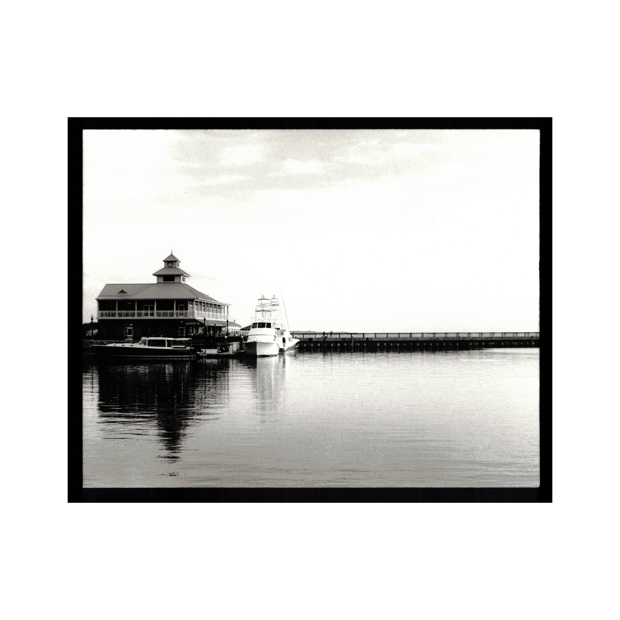 A black and white photo of a marina with two boats docked near a building with a tower, and a long pier extending into the water.