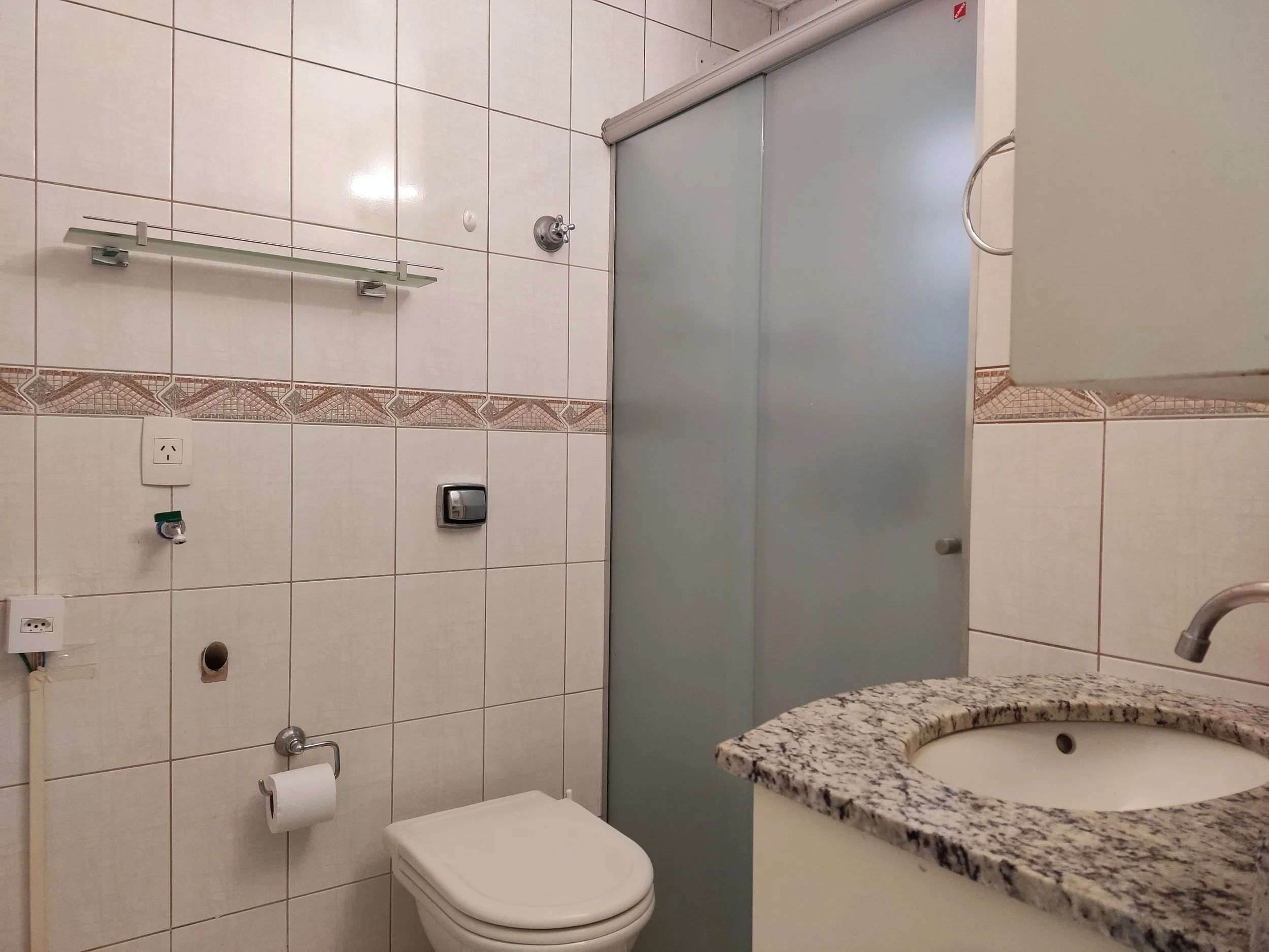 Banheiro com pia de granito, vaso sanitário, ducha e armário de vidro, paredes de azulejo branco com detalhe decorativo em linha na altura média.